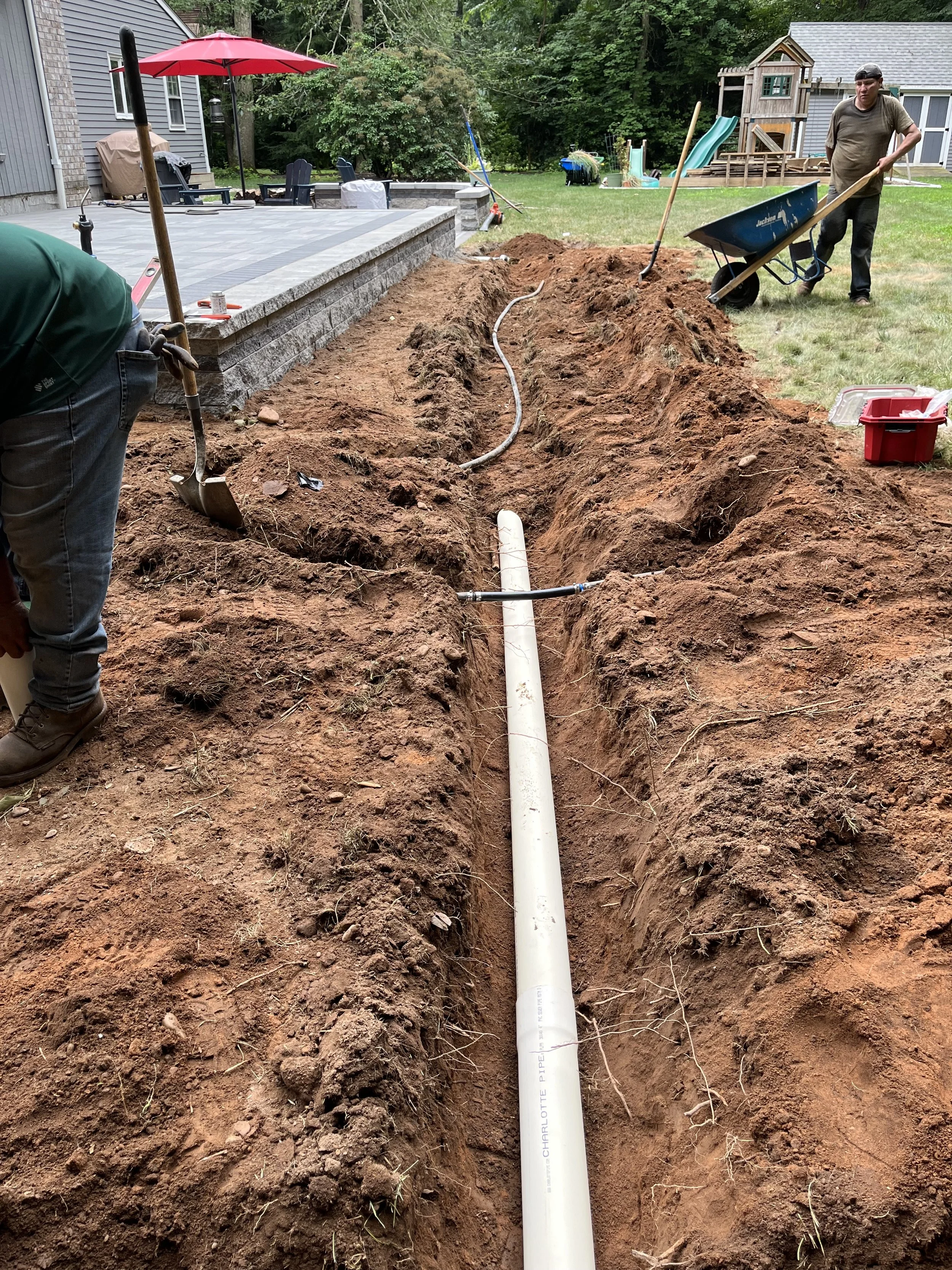 People installing PVC pipes in a trench for underground plumbing or electrical lines in a backyard. The trench runs alongside a new concrete patio and a grassy yard with play equipment.