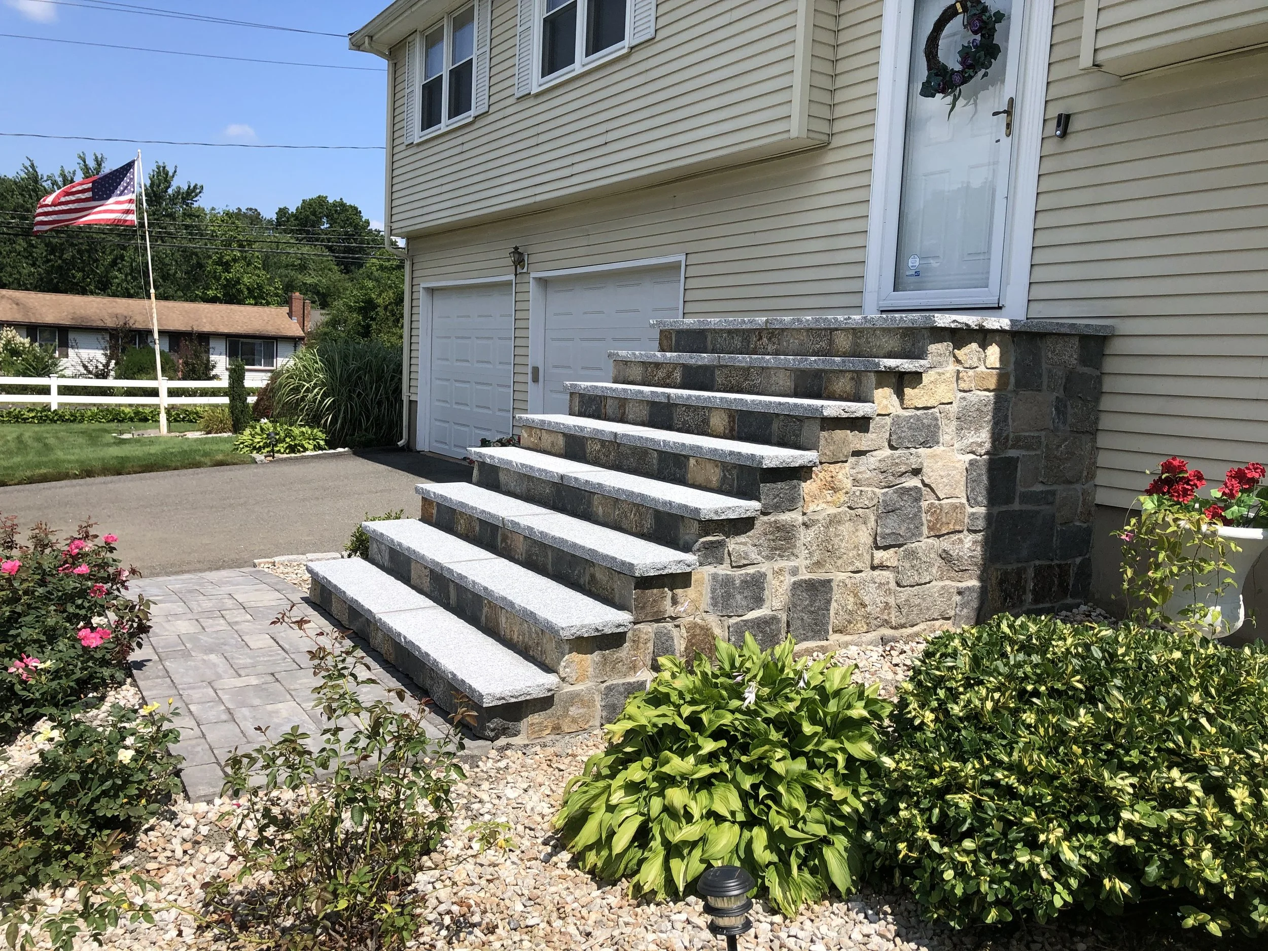 Stone steps leading up to the front door of a beige house with a wreath on the door. The house has a garage and a driveway, with flowers and bushes in the garden. An American flag is visible in the background.