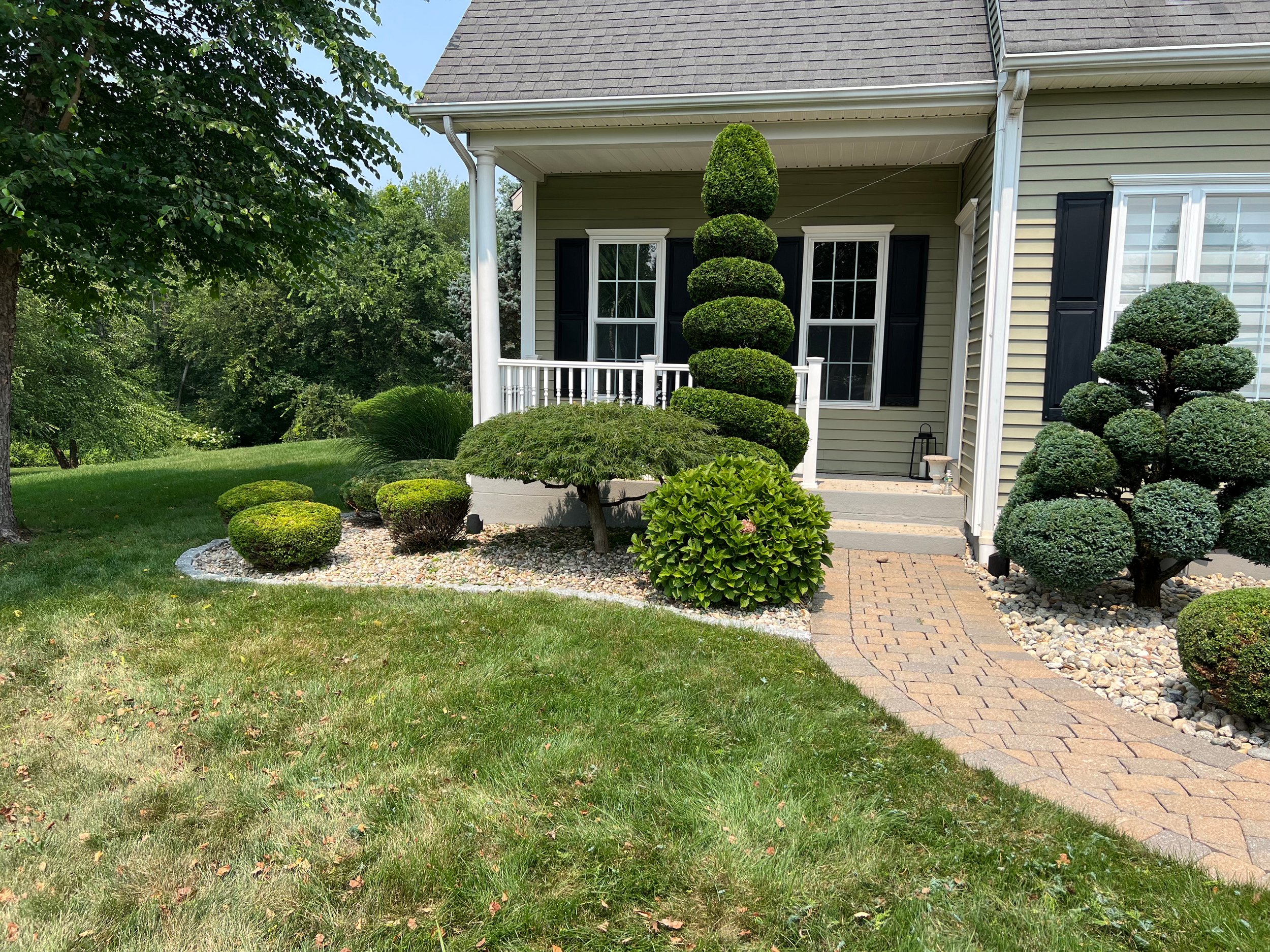 Neatly landscaped front yard with trimmed bushes and a winding brick walkway leading to the porch of a house with beige siding and black shutters.