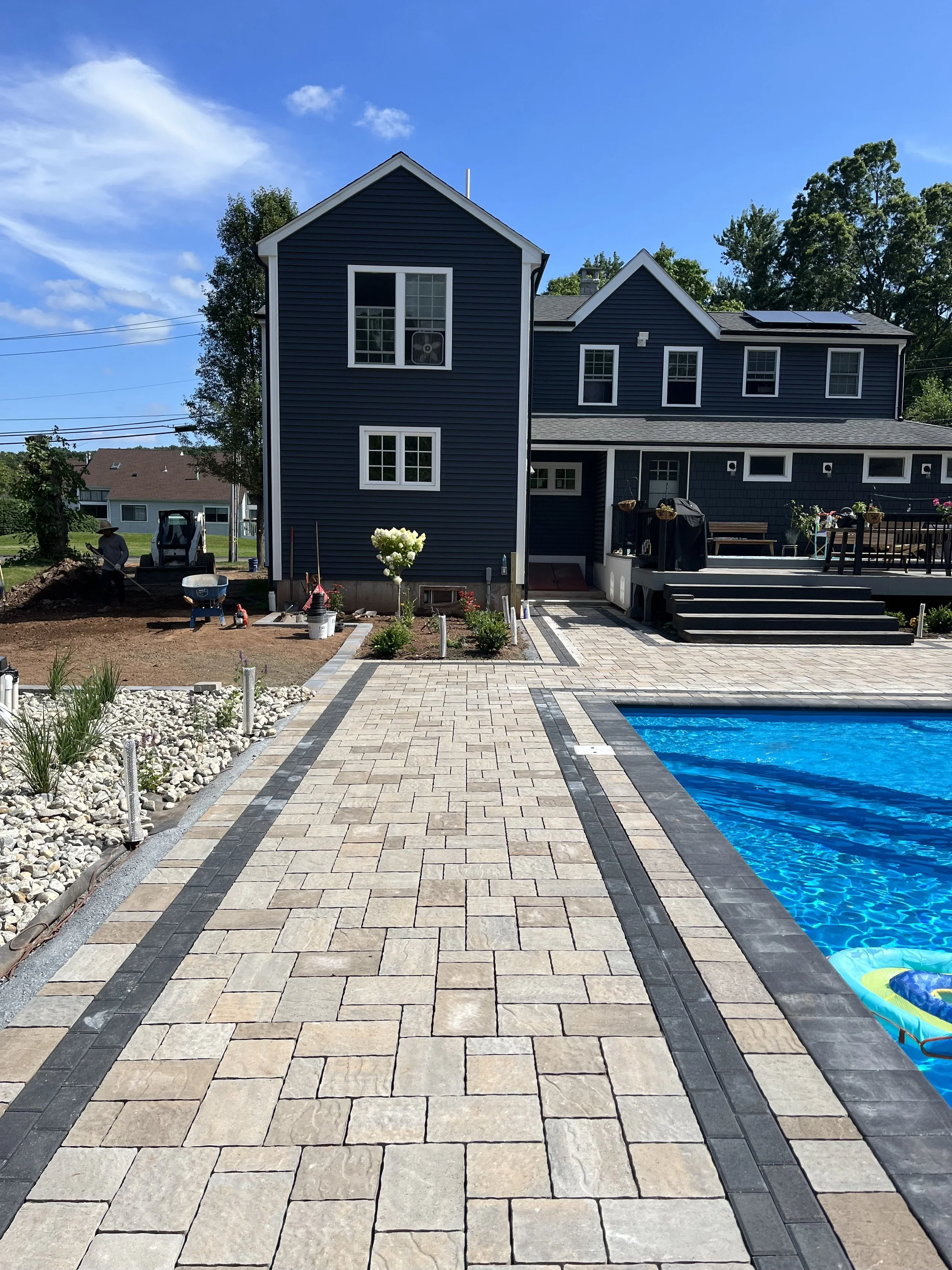 New backyard with a paved patio, swimming pool, and a house with dark blue siding and white trim in sunny weather.
