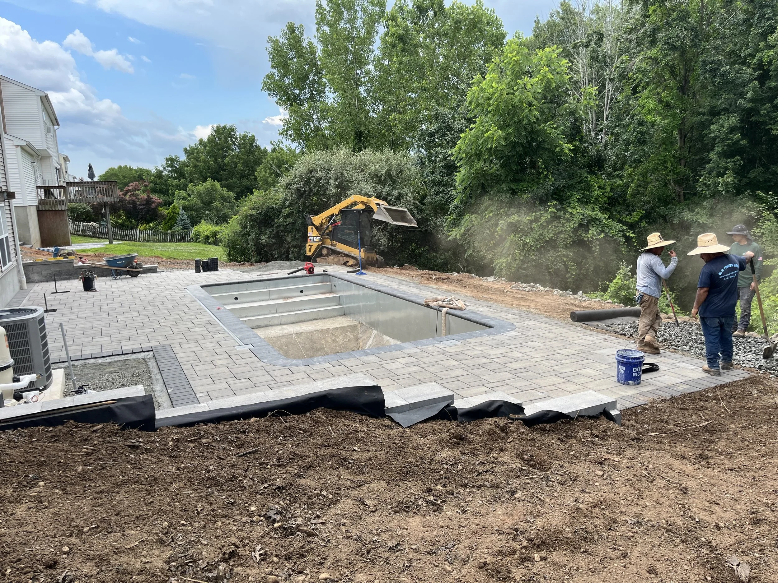 Construction workers installing pavers around a swimming pool with a backyard and trees in the background.