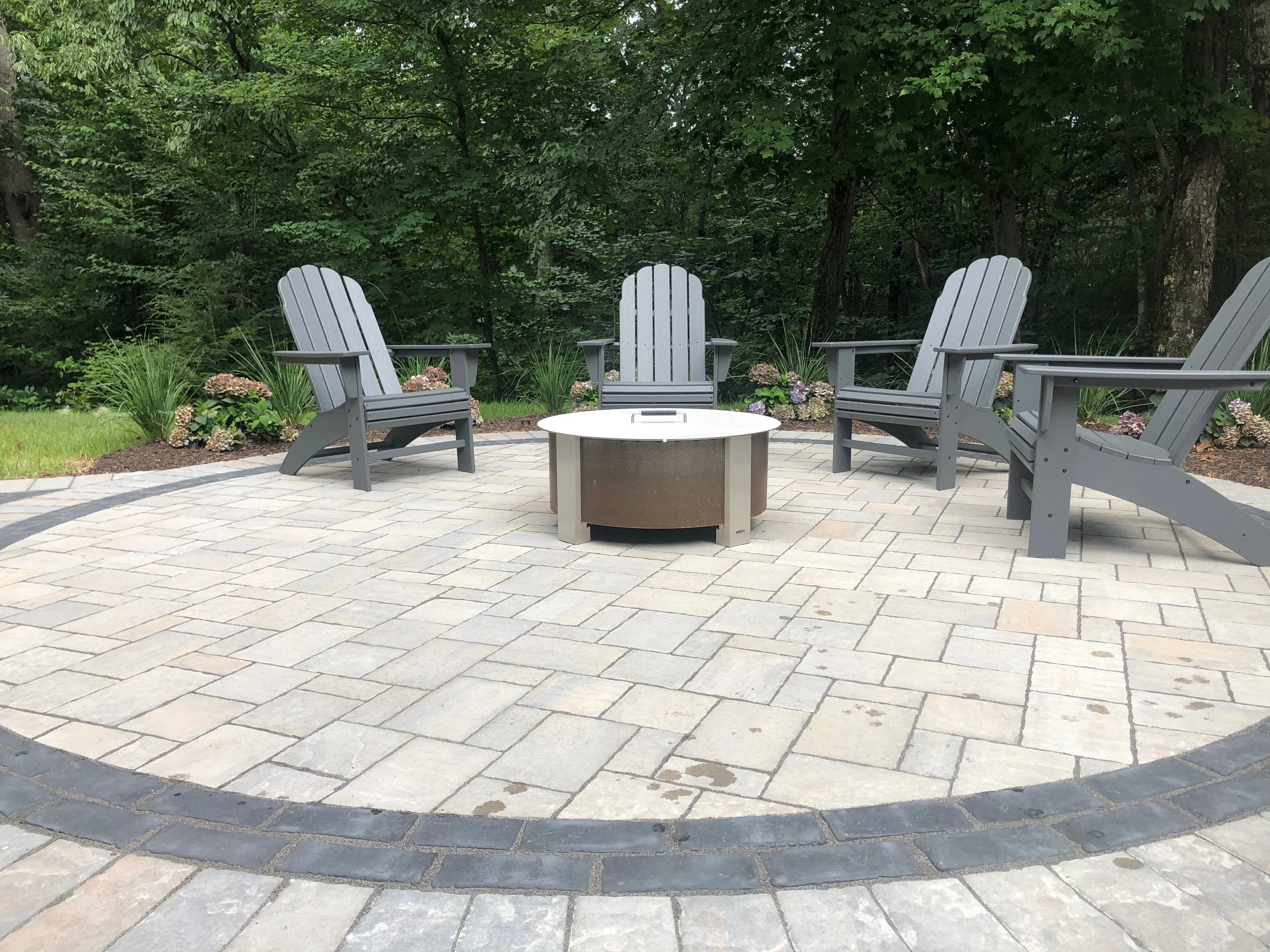 A backyard fire pit area with four gray Adirondack chairs arranged around a steel fire pit on a stone patio