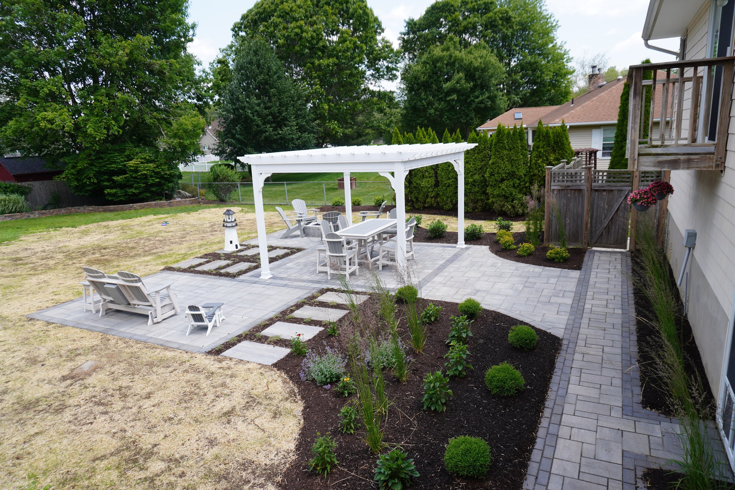 Backyard patio with white outdoor furniture, a pergola, garden beds with bushes and flowers, a lighthouse decoration, and a wooden fence with gates, surrounded by trees.
