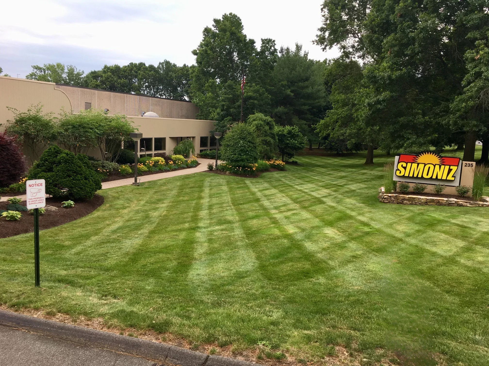 A neatly maintained grassy lawn with striped mowing pattern, landscaped bushes and flowers near a building, and a large sign that reads "SIMONZ" in yellow and black letters, surrounded by small plants and a stone border.