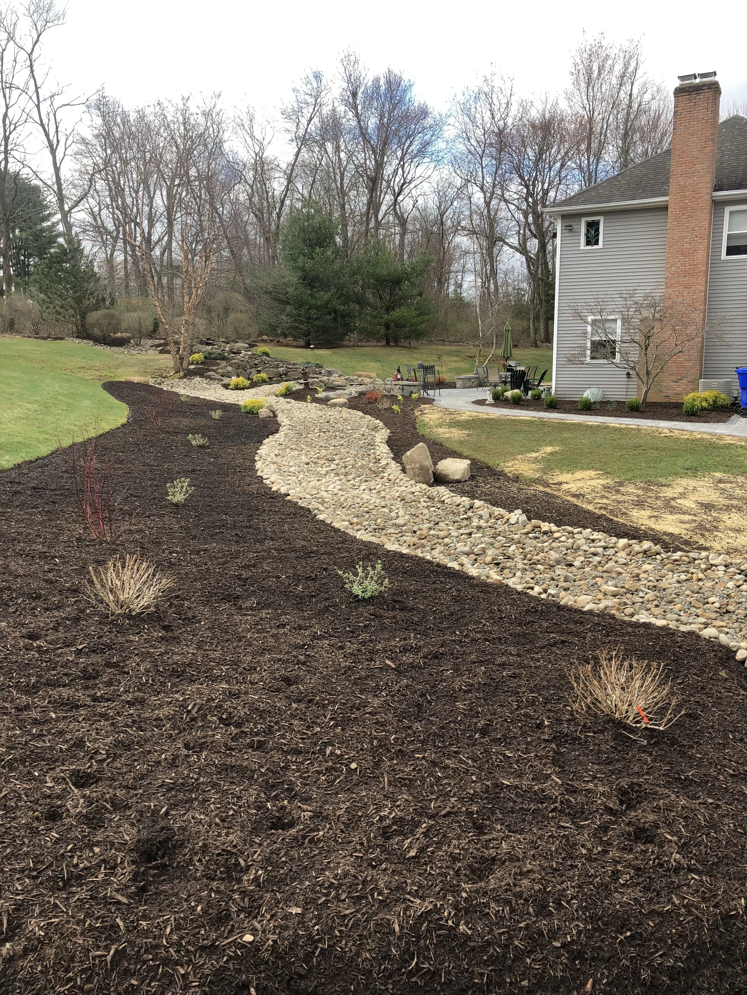 A winding stone pathway through a landscaped backyard with young plants and a lawn, adjacent to a gray house with a brick chimney.