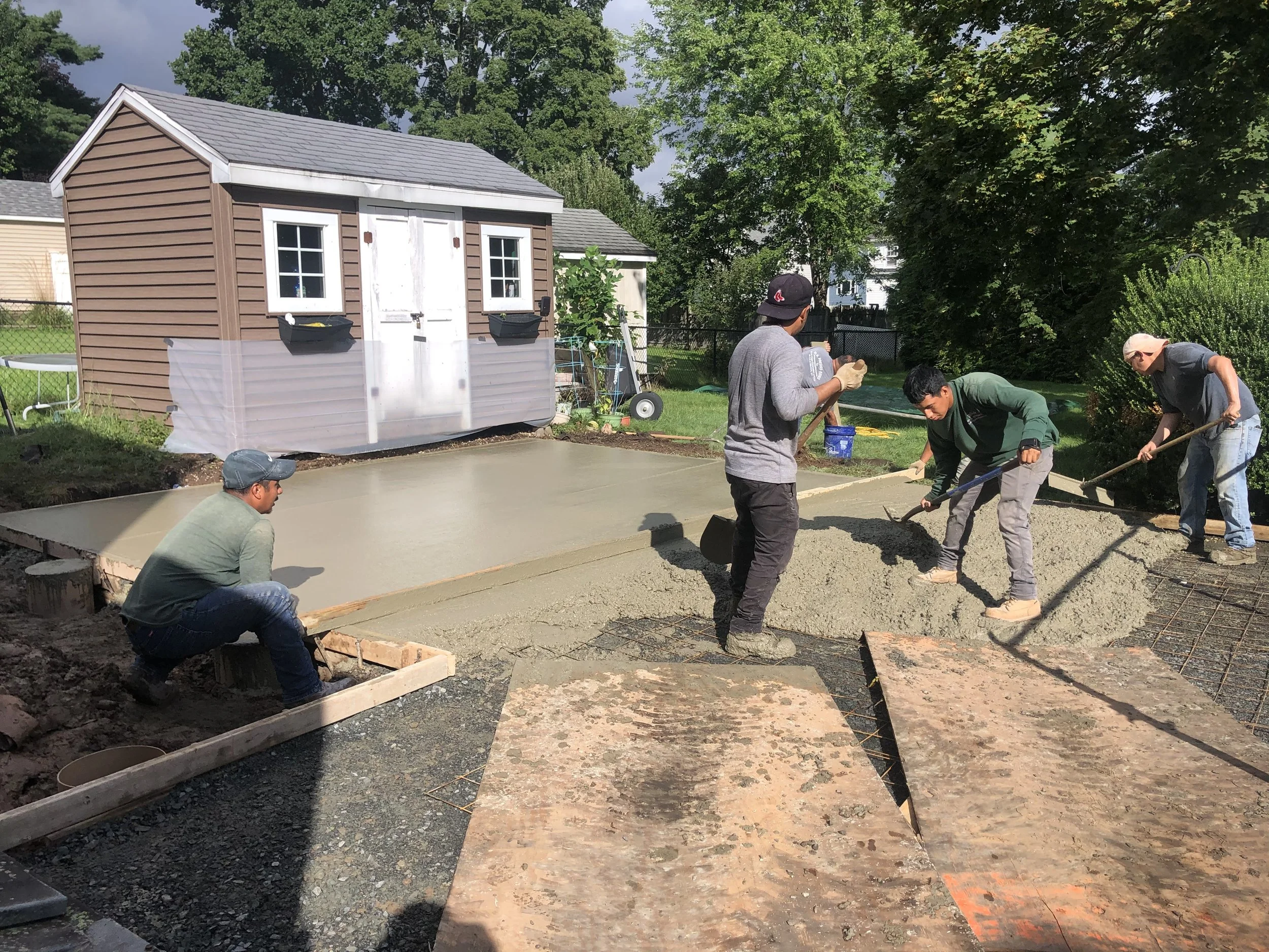 People working on a concrete slab in a backyard, with a small shed in the background and trees surrounding the area.