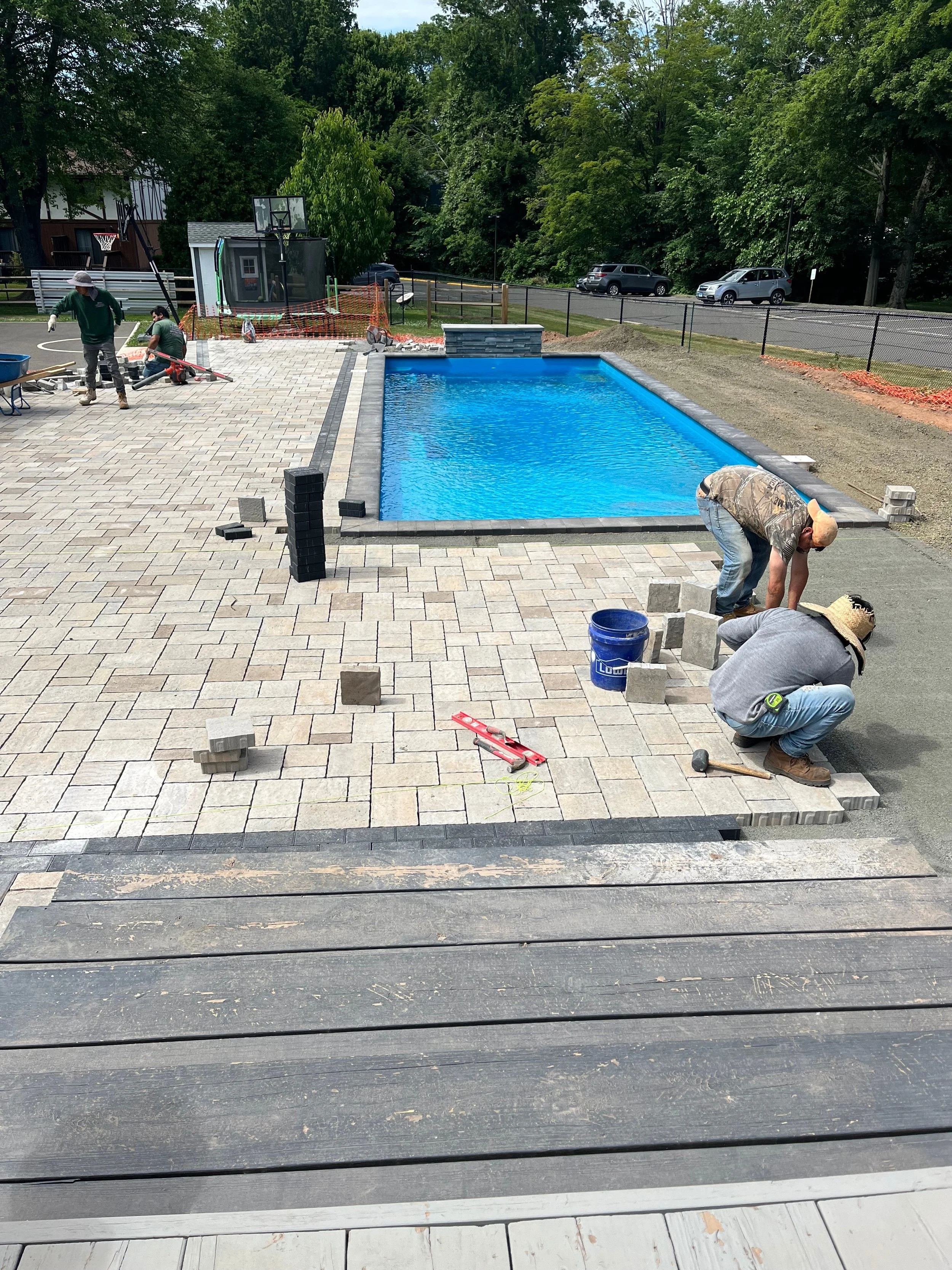 Construction workers installing a brick patio around a backyard swimming pool, with some workers laying bricks and others working nearby.