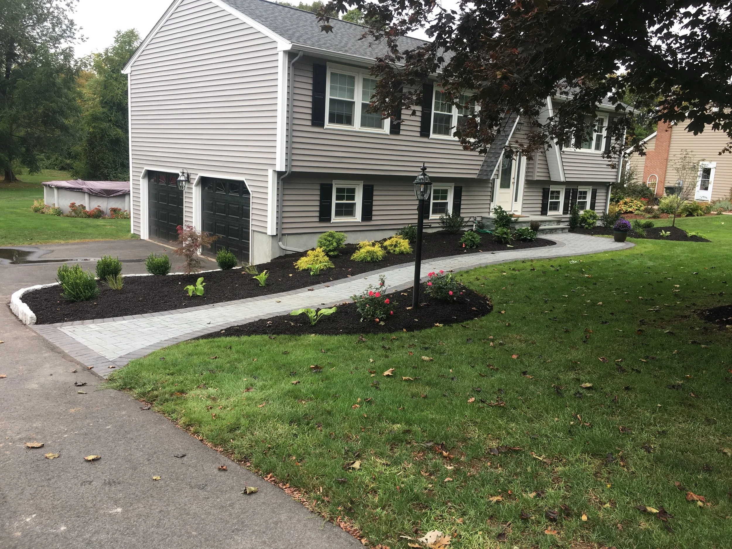 Front yard with a paved walkway, landscaped with small shrubs, flowers, and mulch, leading to a gray house with black shutters and garage doors, surrounded by green grass and trees.