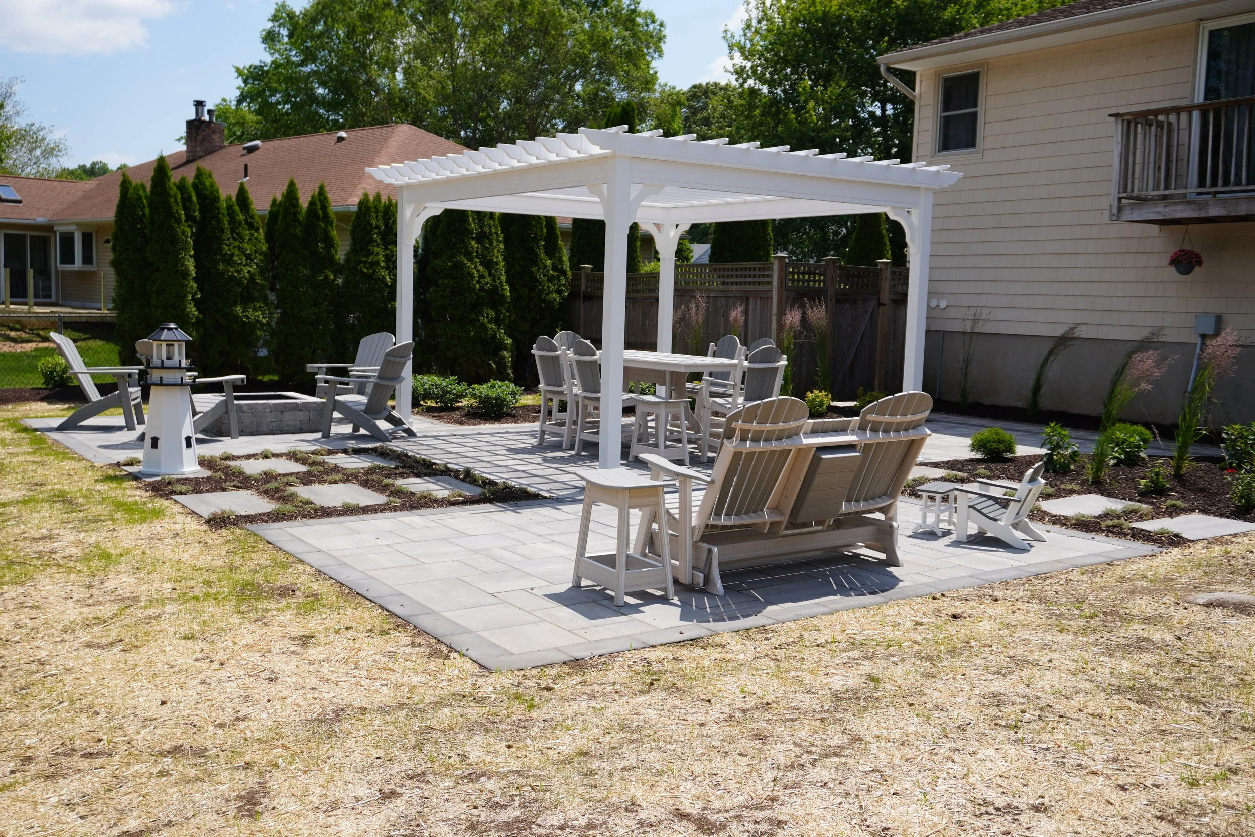 Backyard patio with a white pergola, outdoor furniture including chairs, a loveseat, a table, a small white bench, and a lighthouse-shaped lantern. Surrounding the patio are greenery and neighboring houses.