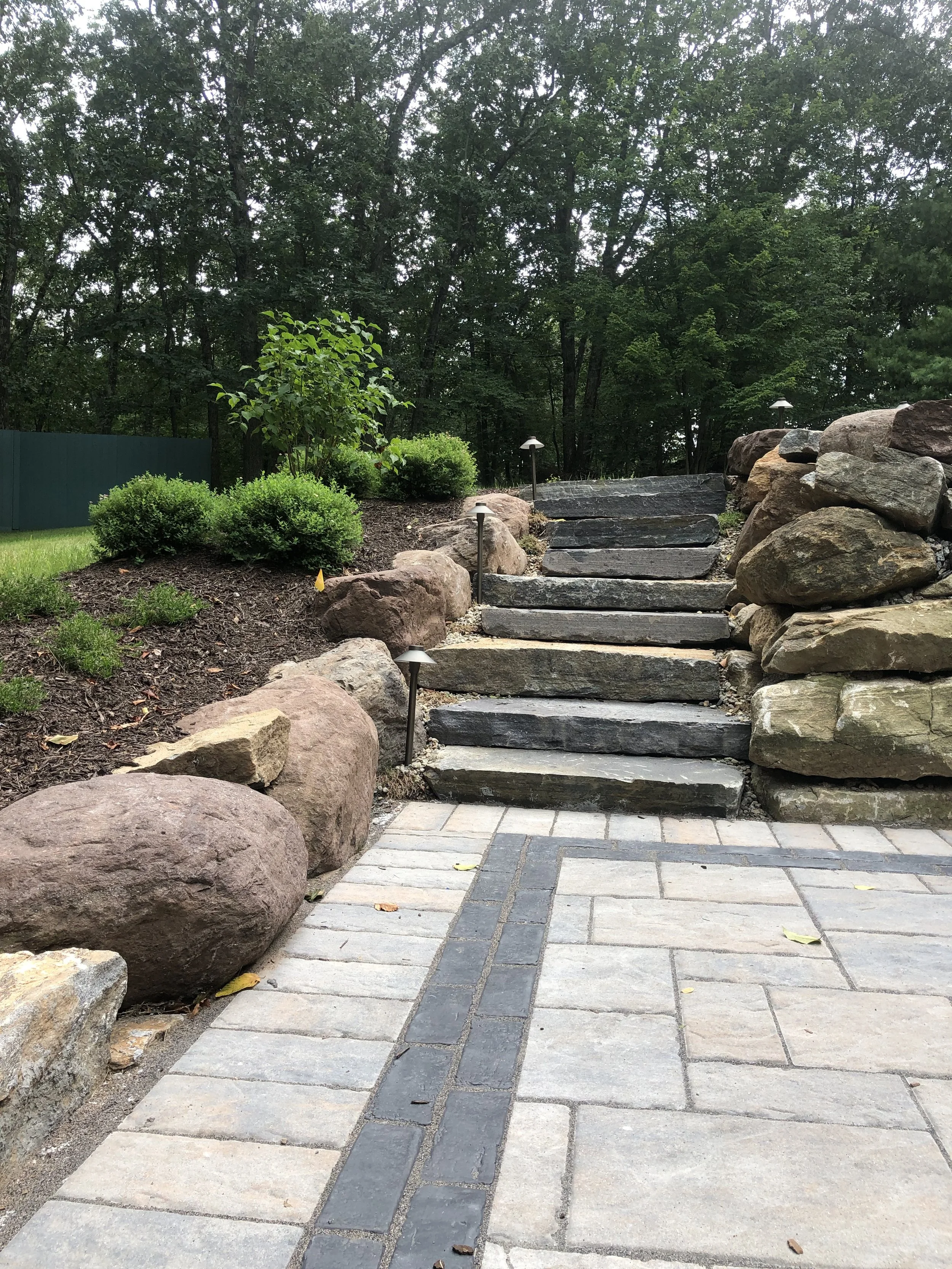 A landscaped backyard with a stone pathway leading to stone steps, surrounded by shrubs and boulders, with a wooded area in the background.