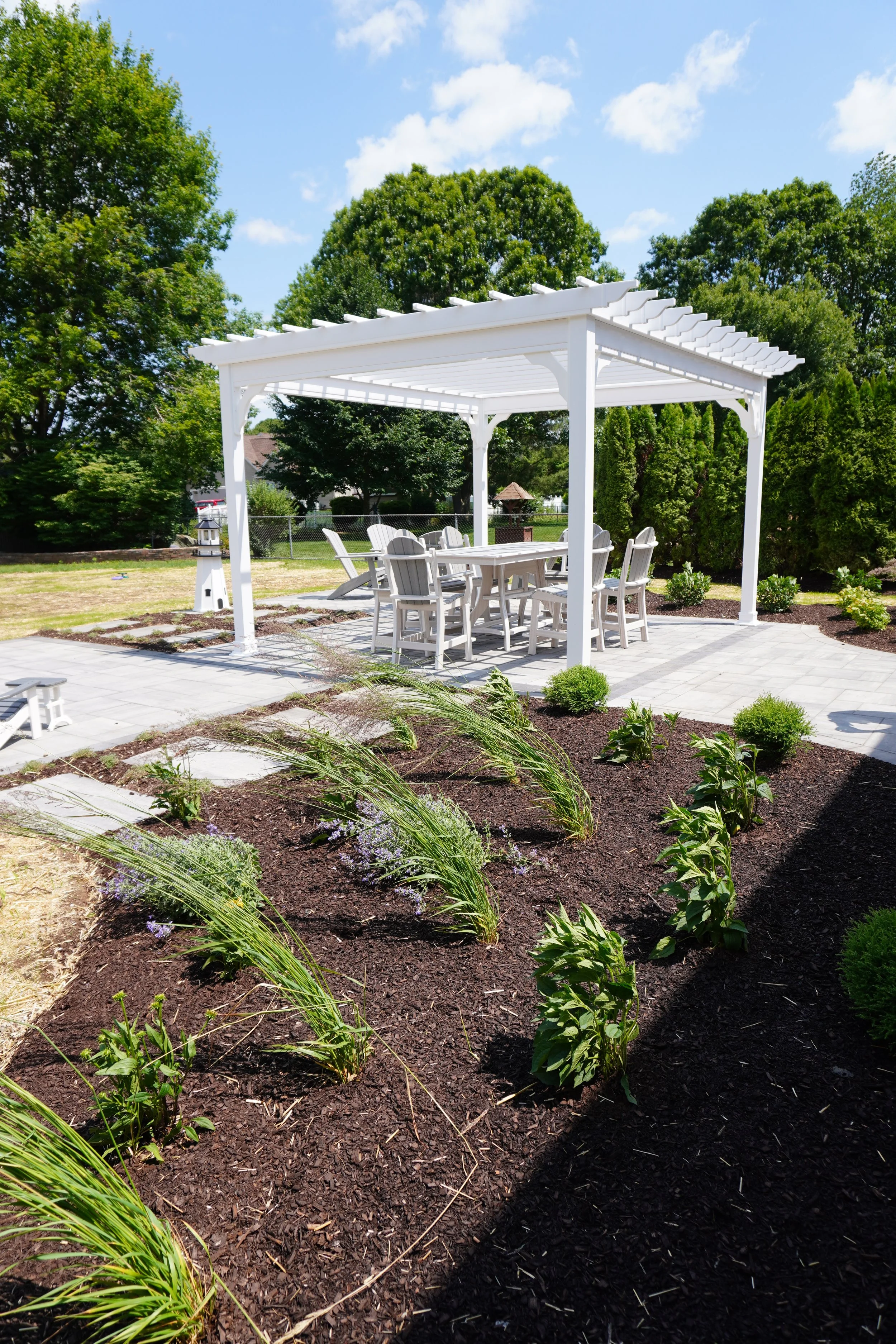 A white pergola with patio furniture underneath, surrounding a garden with mulched flower beds, greenery, and a sunny blue sky.