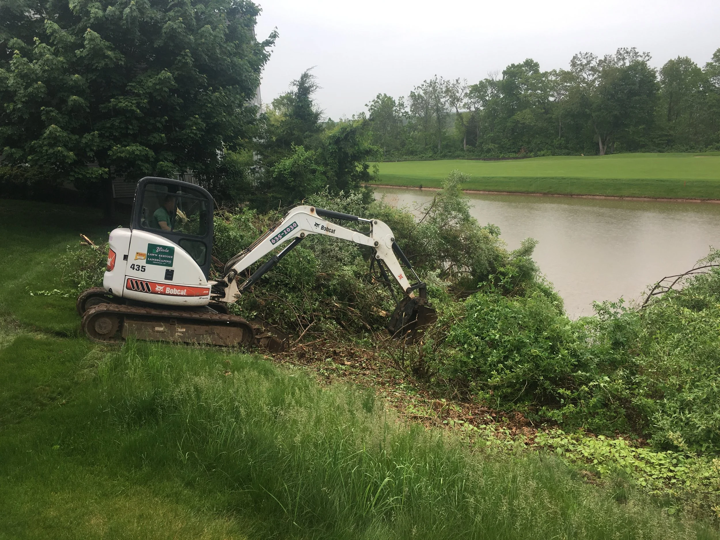A small excavator removing bushes by a pond on a grassy area, with trees and a golf course in the background.