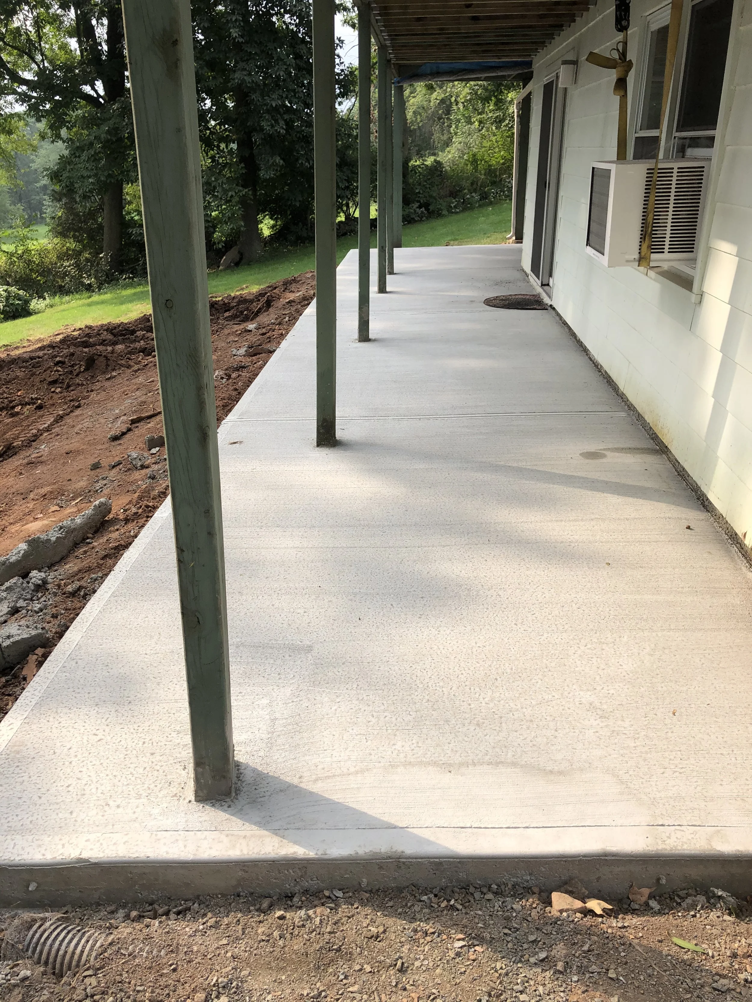 Newly poured concrete patio on a house with a wrapped-around porch. Support posts are in place, and an air conditioning unit is mounted on the wall. The yard has green grass and trees, with dirt along the edge of the patio.