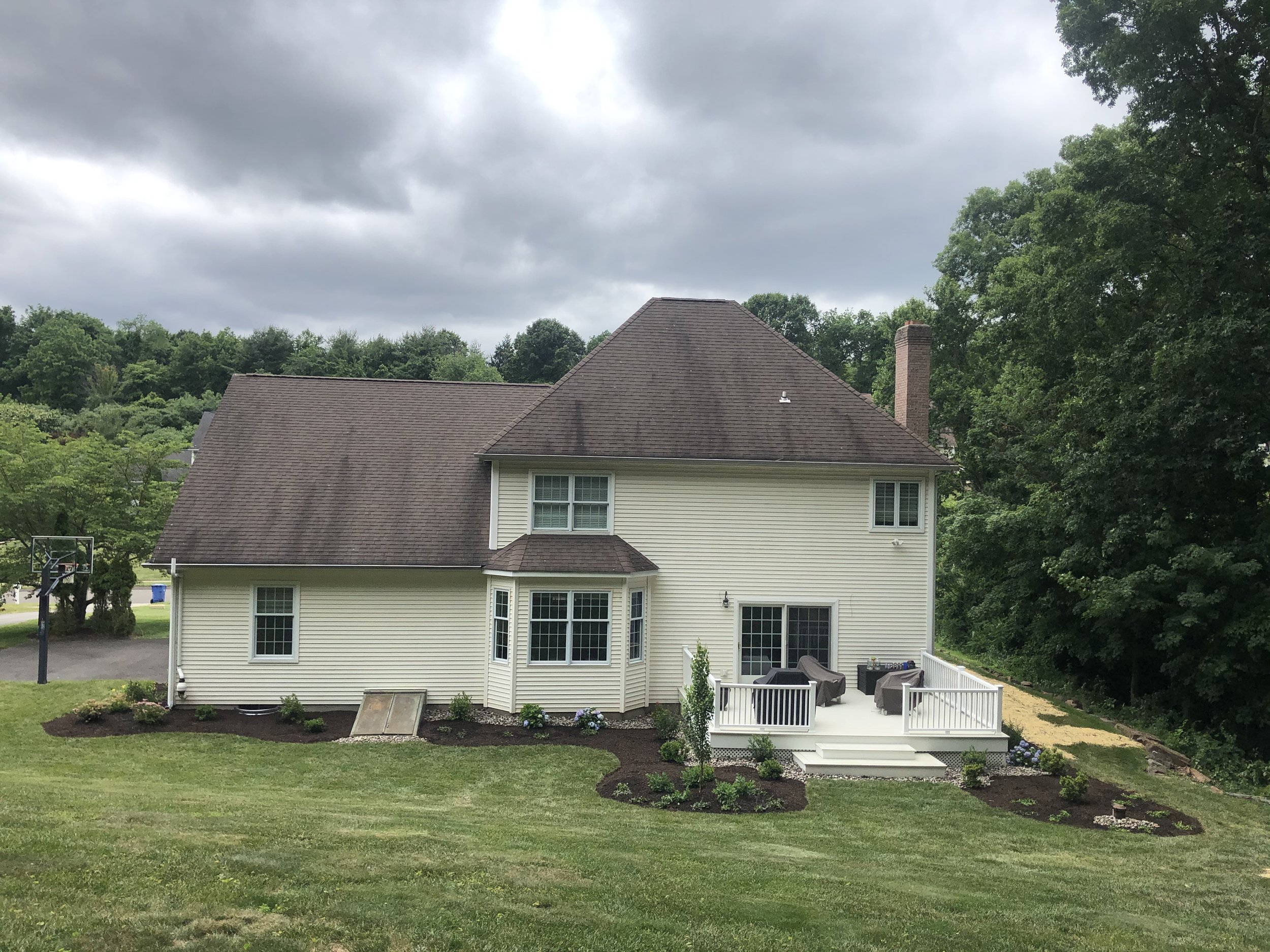 Back view of a two-story beige house with a brown roof, white porch with outdoor furniture, surrounded by a landscaped yard with flowers