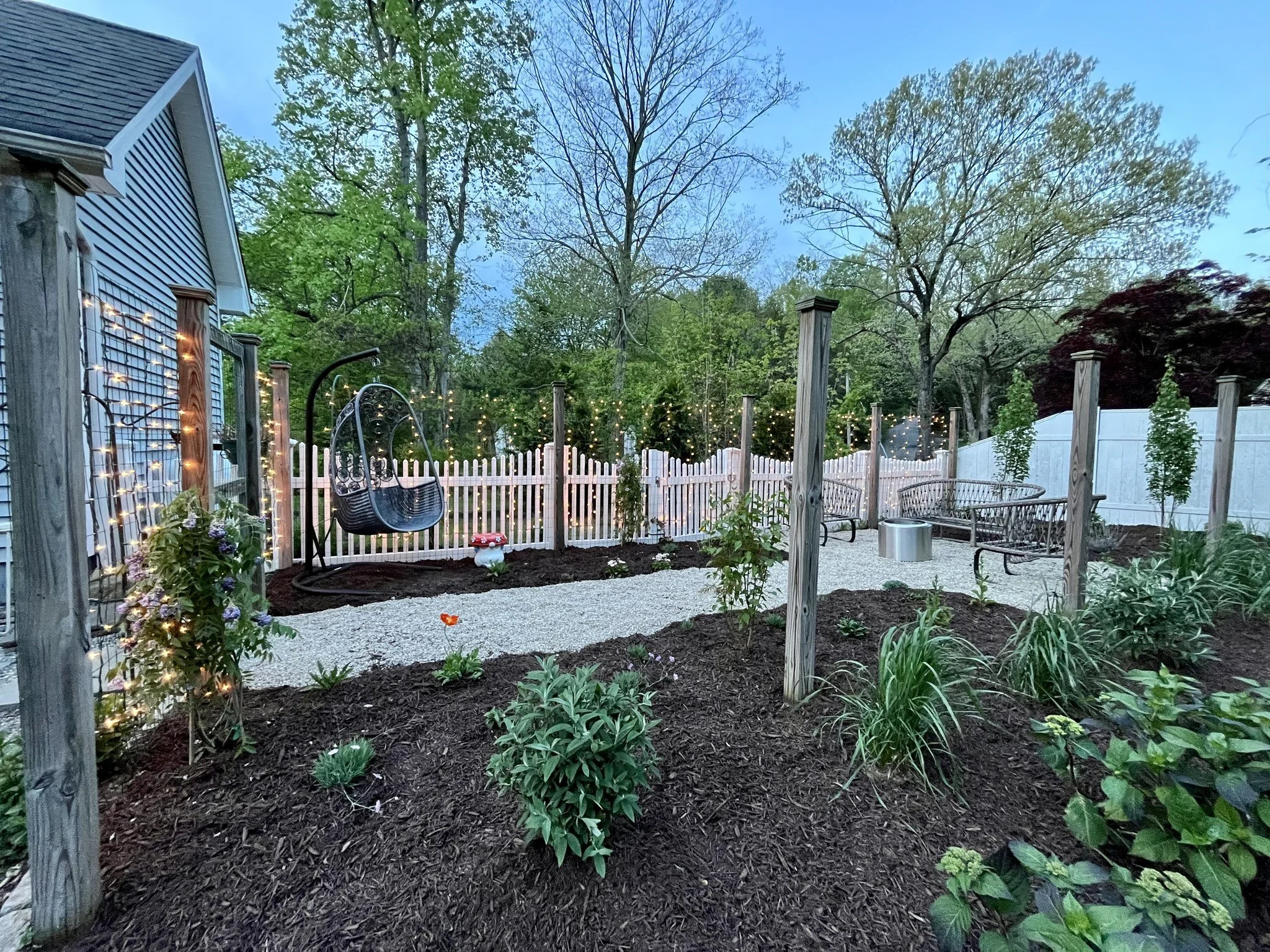 A backyard garden at dusk with a white picket fence, string lights, benches, swing, potted plants, and trees in the background.