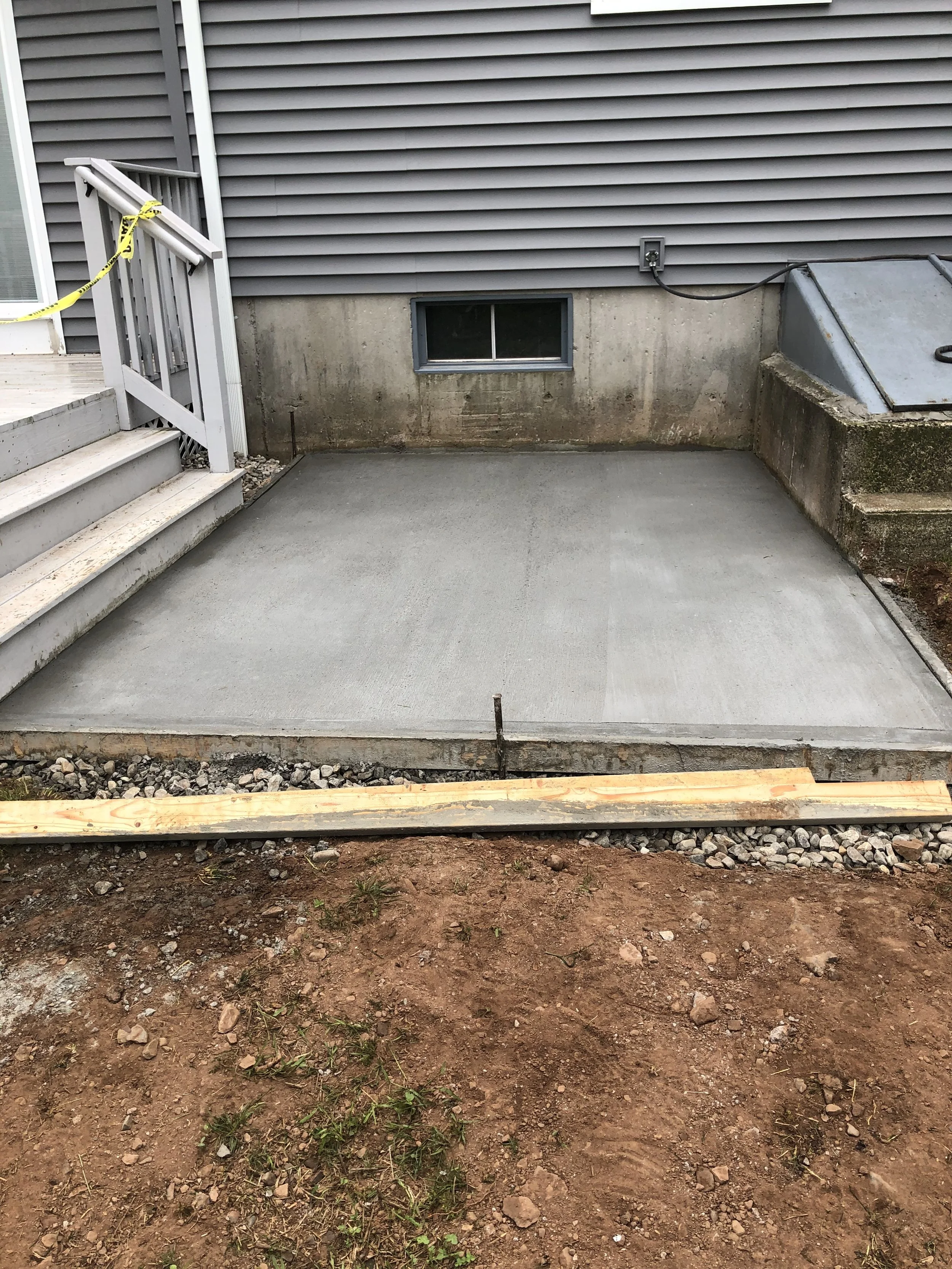 Freshly poured concrete pad next to a house with grey siding, a small basement window, and a set of stairs leading to a deck, with construction materials and dirt in the foreground.