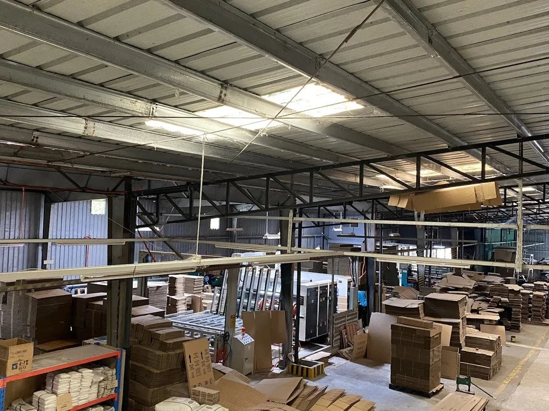 Warehouse with stacked cardboard boxes, packaging materials, and industrial equipment, under a metal roof with skylights.