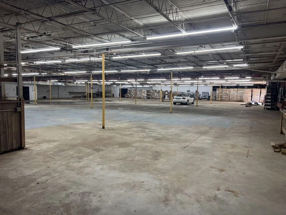 An empty parking garage with concrete floors, yellow support poles, fluorescent lighting, and a few parked cars and motorcycles in the distance.