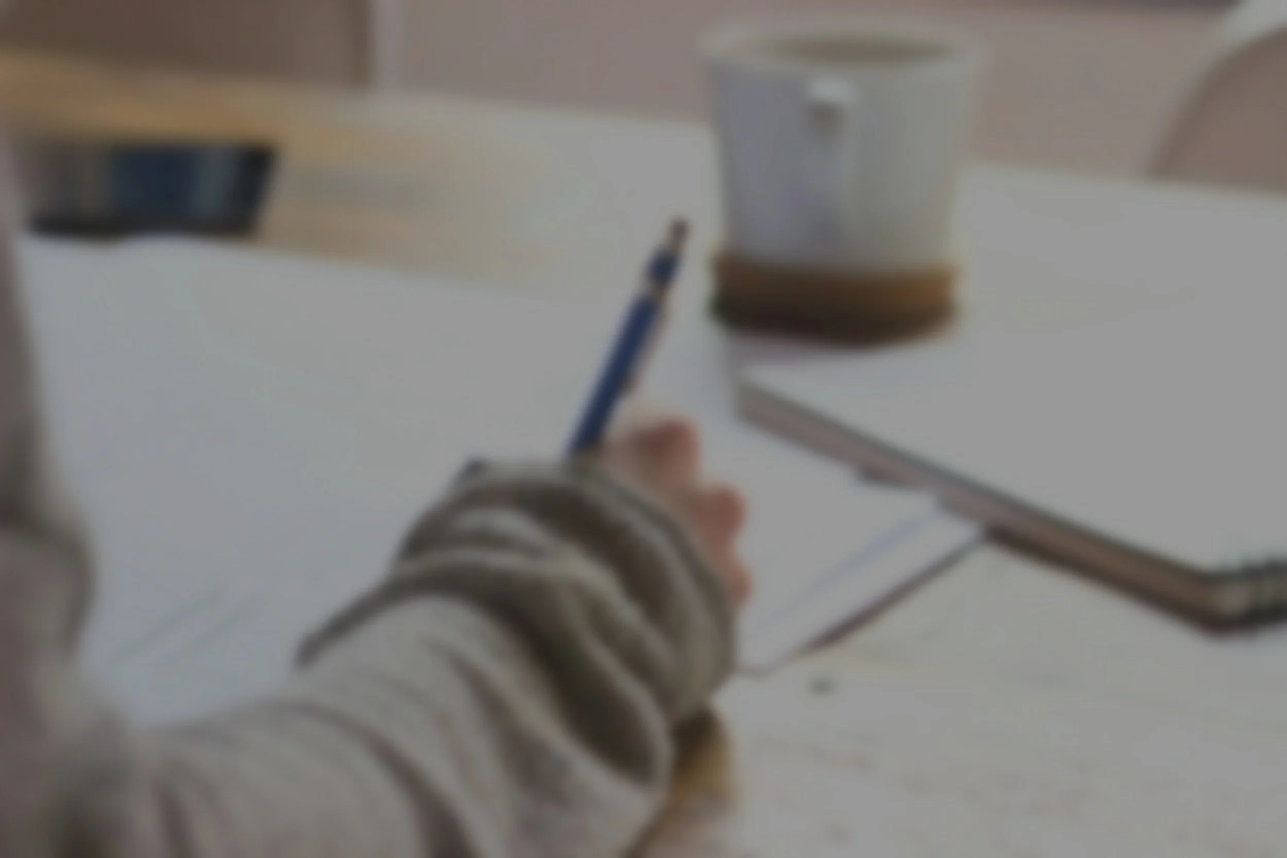 A person with a striped sweater writing on papers with a pen. A coffee cup and notebook on a wooden table.