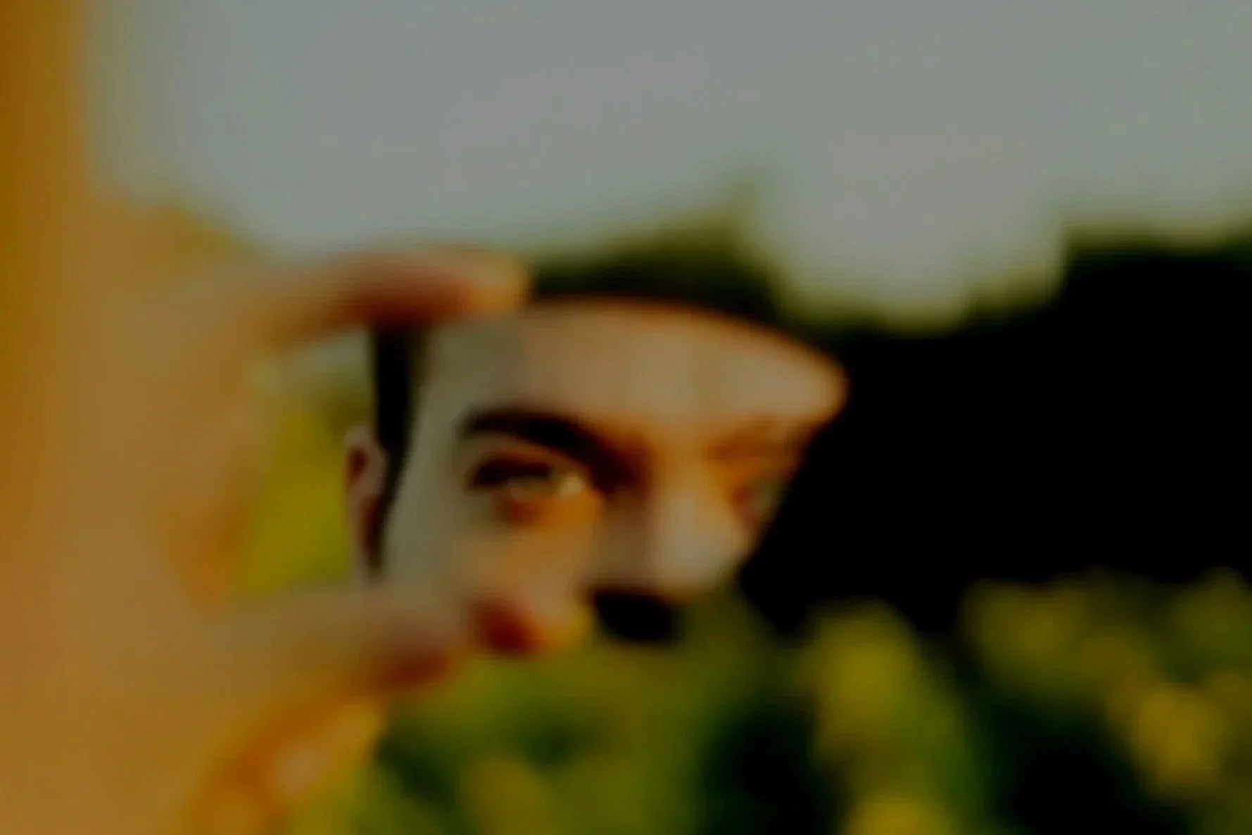 A person with yellow-green eyes looking through a small opening in a plant or bush, with sunlight illuminating parts of their face.