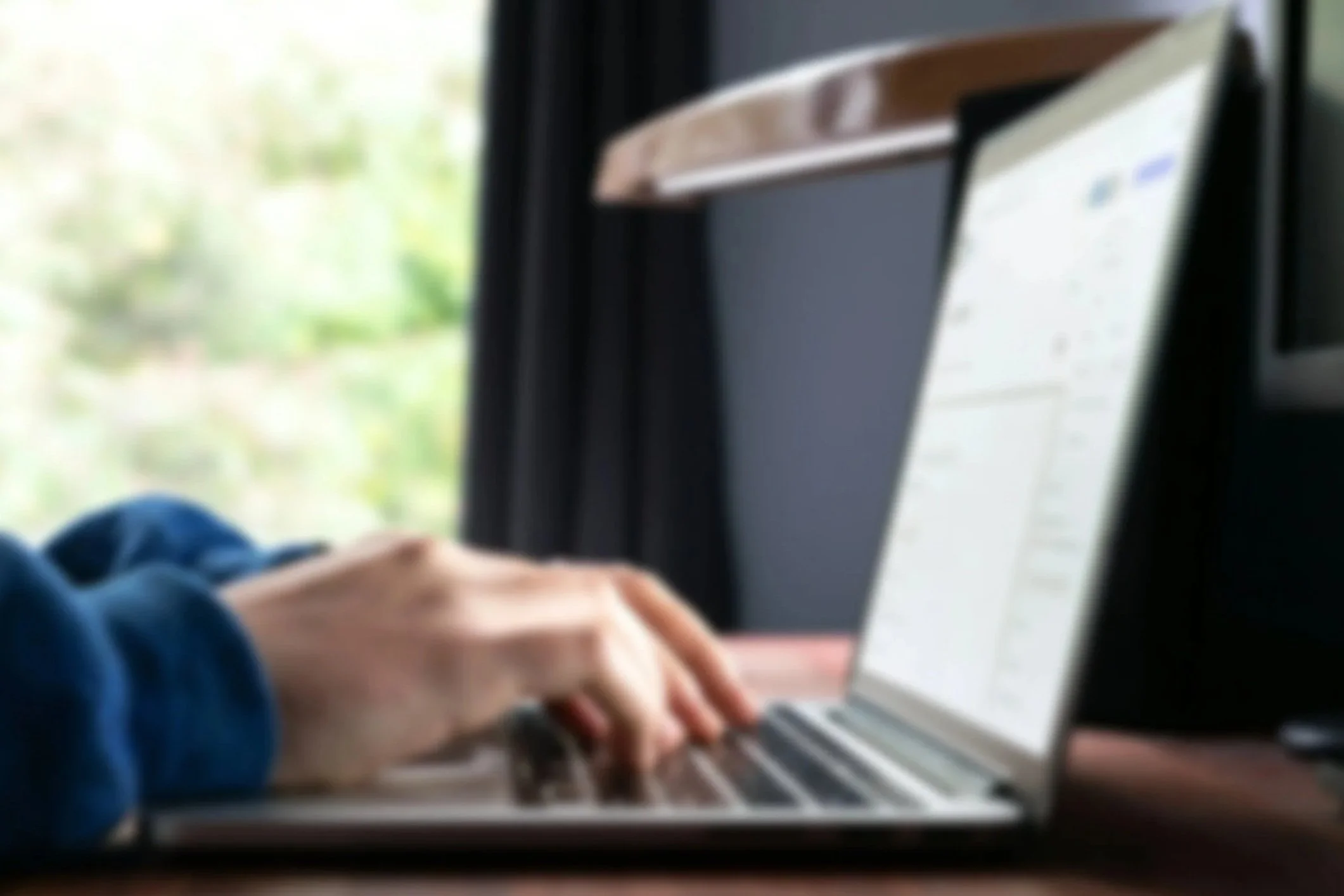 Person wearing a blue jacket typing on a laptop with a blurred window view in the background.