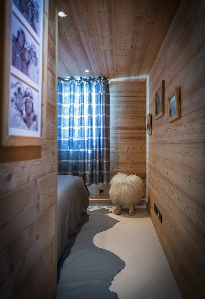 Une petite pièce en bois avec un lit, une fenêtre avec des rideaux à carreaux bleus, un tapis blanc, et un fauteuil en forme de mouton en peluche.