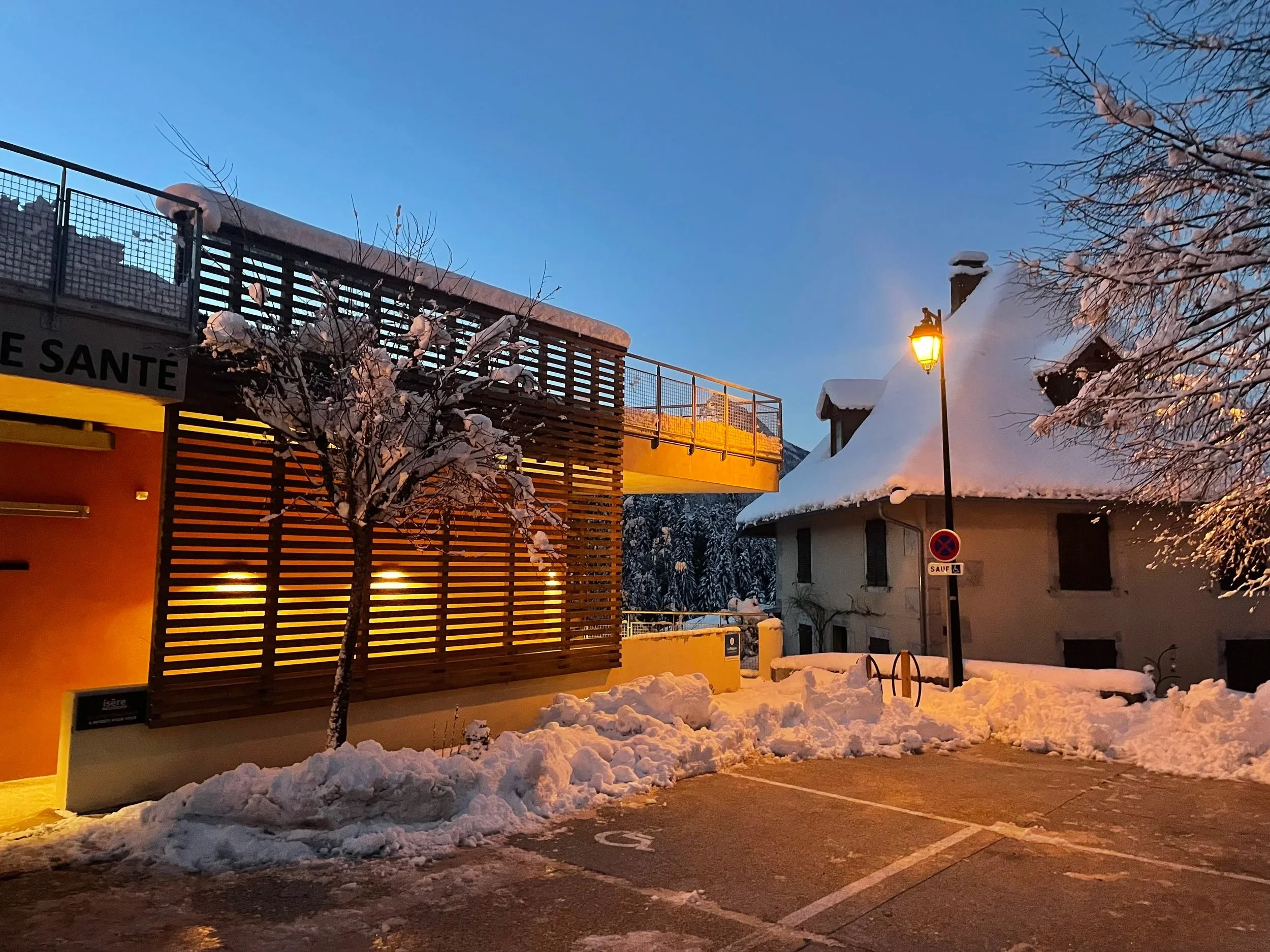 Une scène de nuit enneigée avec une maison et une église, éclairée par un lampadaire, des arbres couverts de neige et un parking partiellement dégagé.