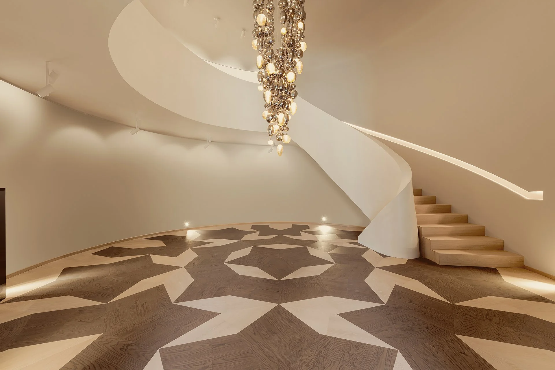 Entrée d'une maison avec un escalier en colimaçon en bois clair, un plafond blanc courbe, un chandelier suspendu en cristal avec lumières chaudes, et un sol en parquet avec un motif géométrique en deux tons.