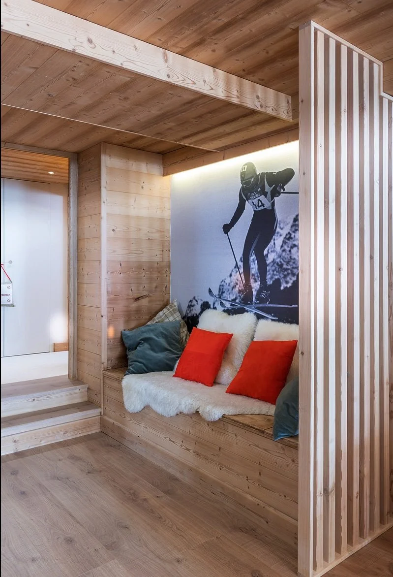 Coin salon avec canapés en bois, coussins rouges et gris, tapis blanc, mural avec image d'un skieur en montagne.