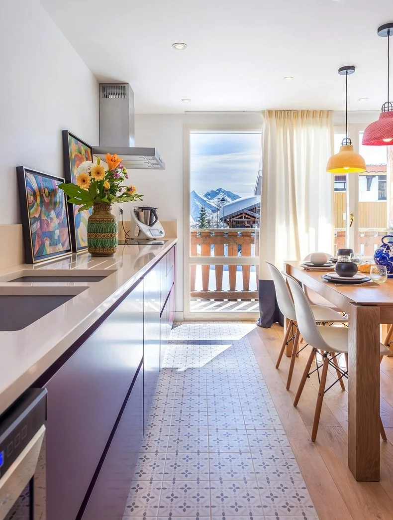 Intérieur d'une cuisine lumineuse avec un balcon donnant sur des montagnes enneigées, un plan de travail avec des fleurs en vase, un robot de cuisine, une table avec des plats et des chaises, et un éclairage suspendu coloré.