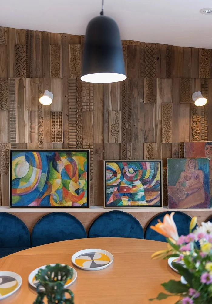 Salle de séjour avec bois mural, lampes encastrées, œuvres d'art abstraites, canapé en velours bleu, table en bois avec vaisselle et fleurs.