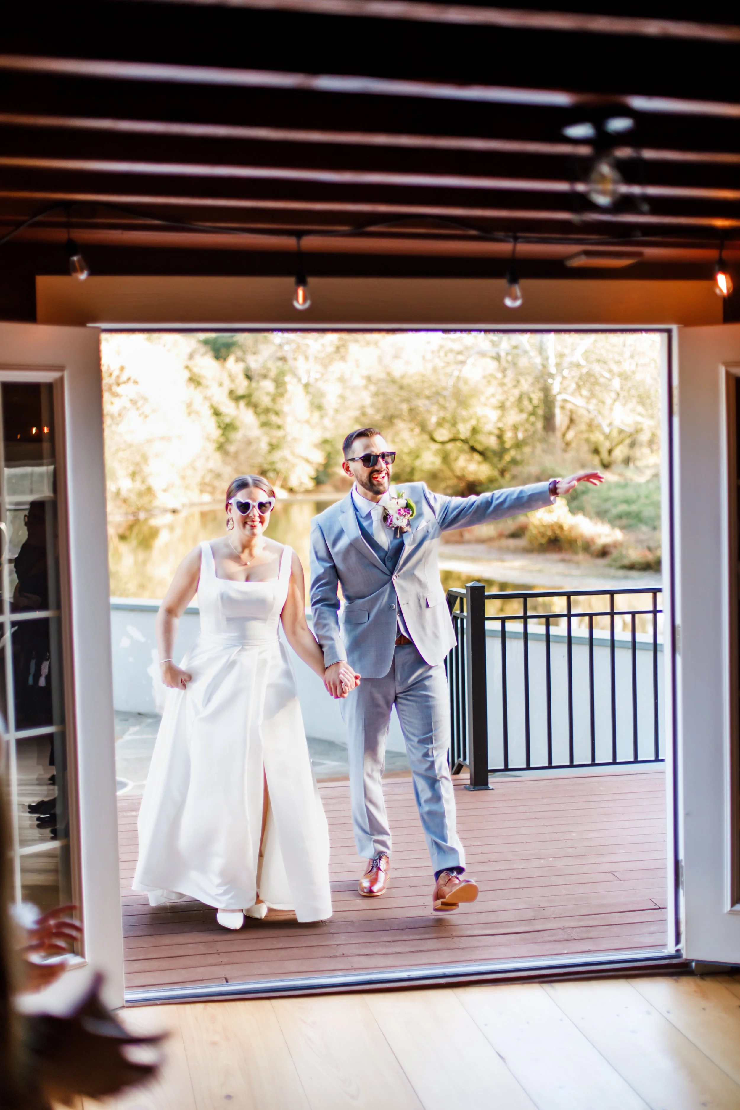A newlywed couple holding hands walking into the wedding venue. The bride is wearing a white wedding gown and sunglasses, and the groom is dressed in a gray suit with sunglasses, pointing outward. The background features trees and a body of water.