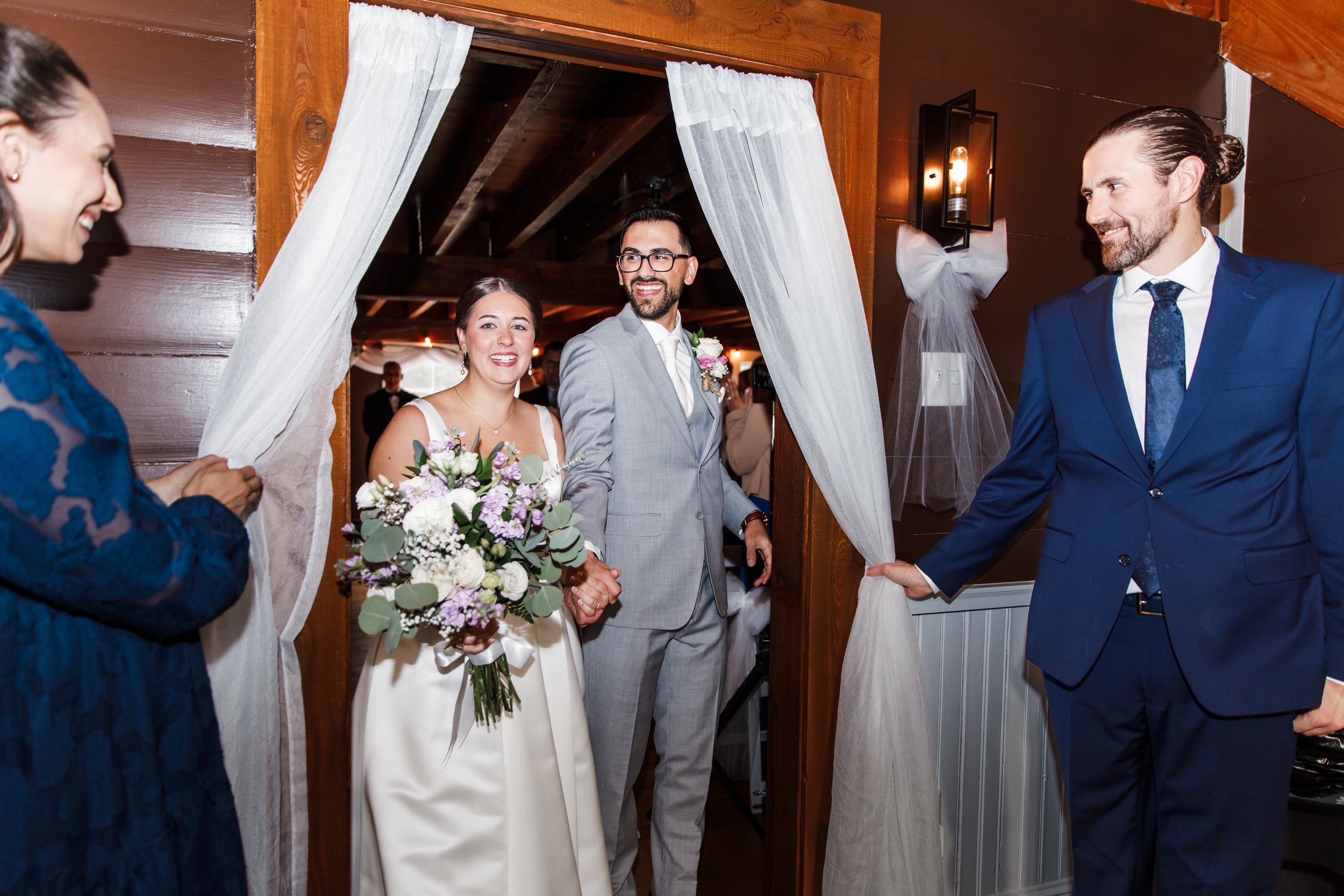 A bride and groom holding hands and smiling as they enter a wedding reception, surrounded by guests in a warmly decorated indoor setting.