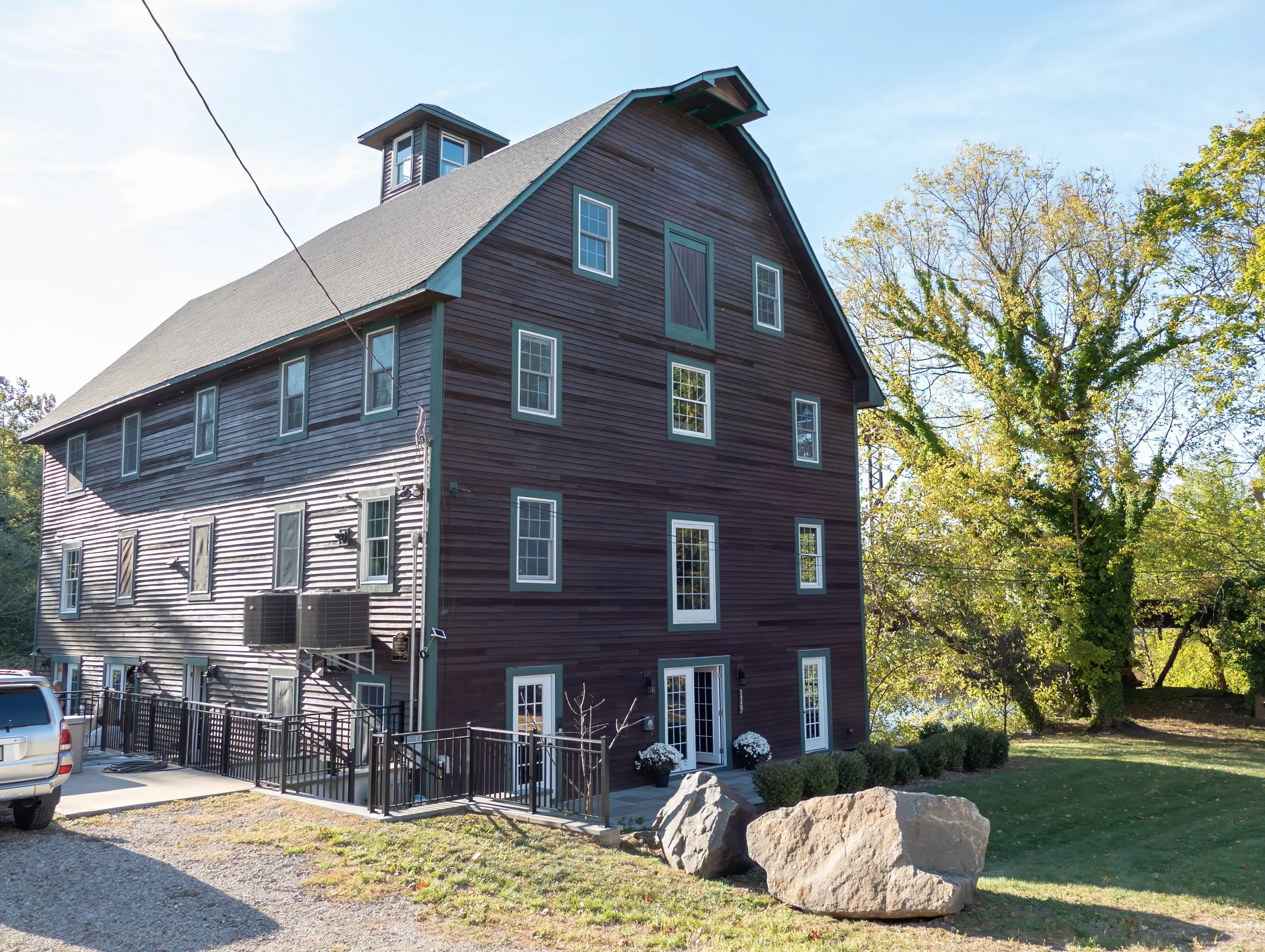 A large wooden multi-story building with a gabled roof, numerous windows, and a small dormer. There is a vehicle parked in front and a black metal railing along the side. Two large rocks are situated on the yard, and there are bushes and tall trees i