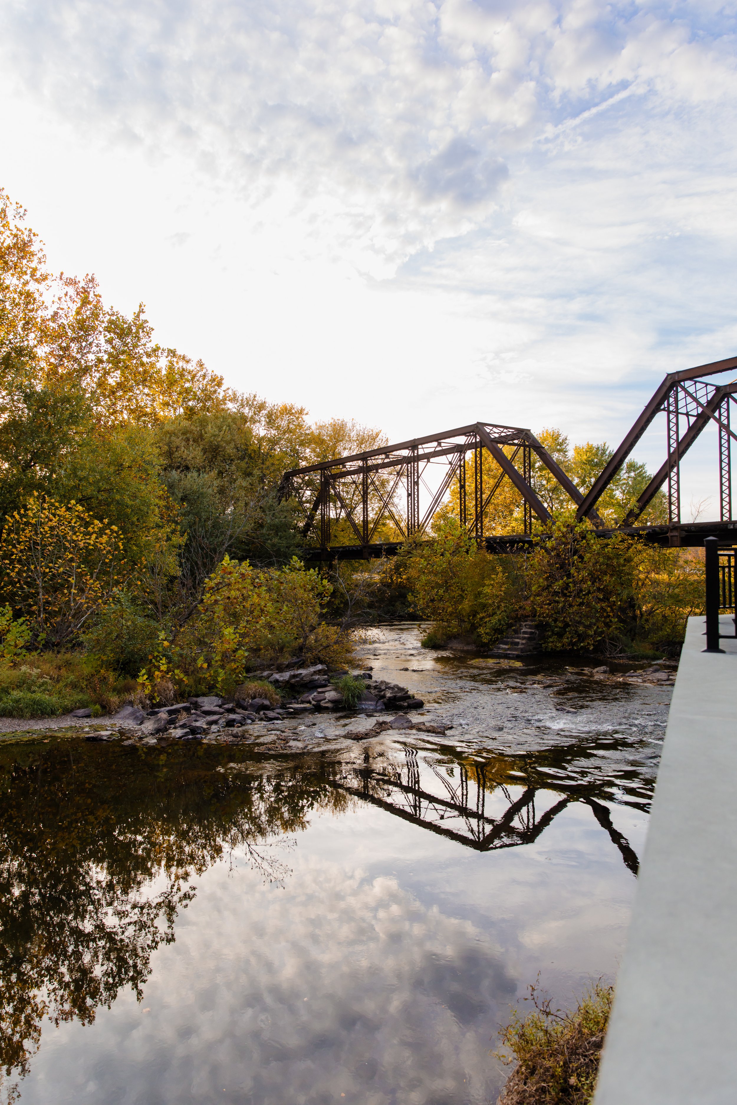 An outdoor scene featuring a rustic metal bridge over the South Branch Raritan River, with trees in autumn foliage on both sides and a partly cloudy sky above.