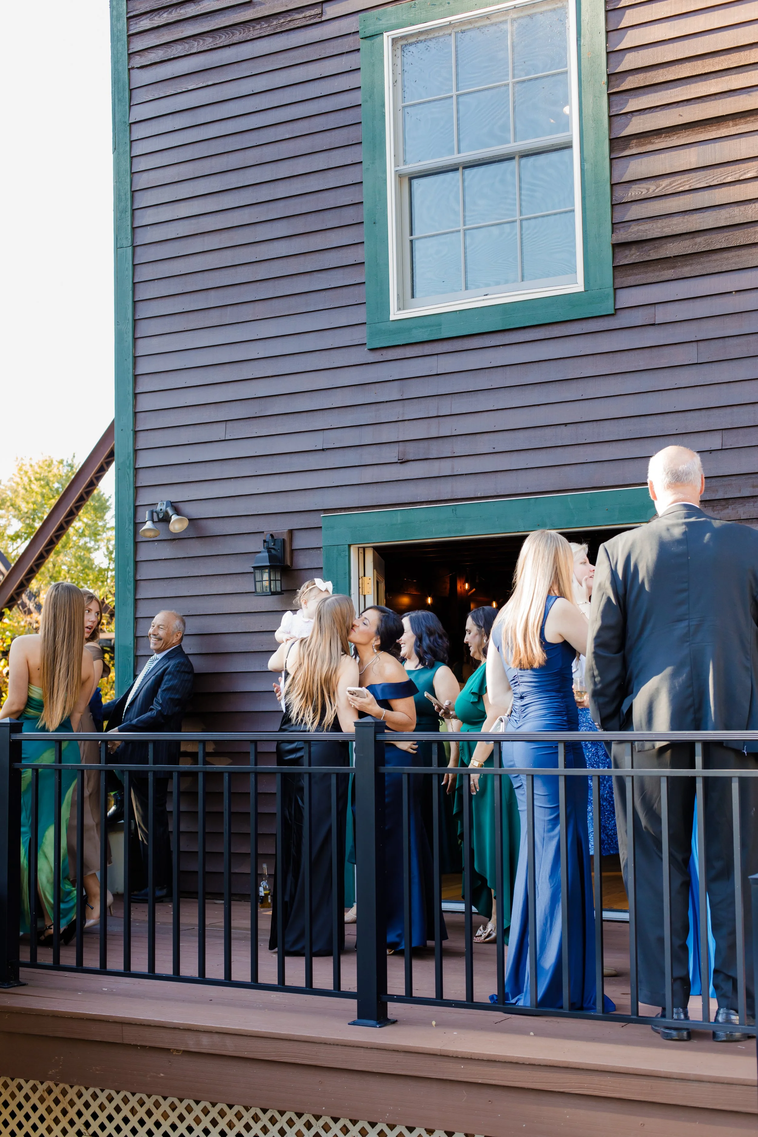 People dressed in formal attire socializing on an outdoor patio at the cocktail hour of a wedding or social event, with a dark wooden building in the background.