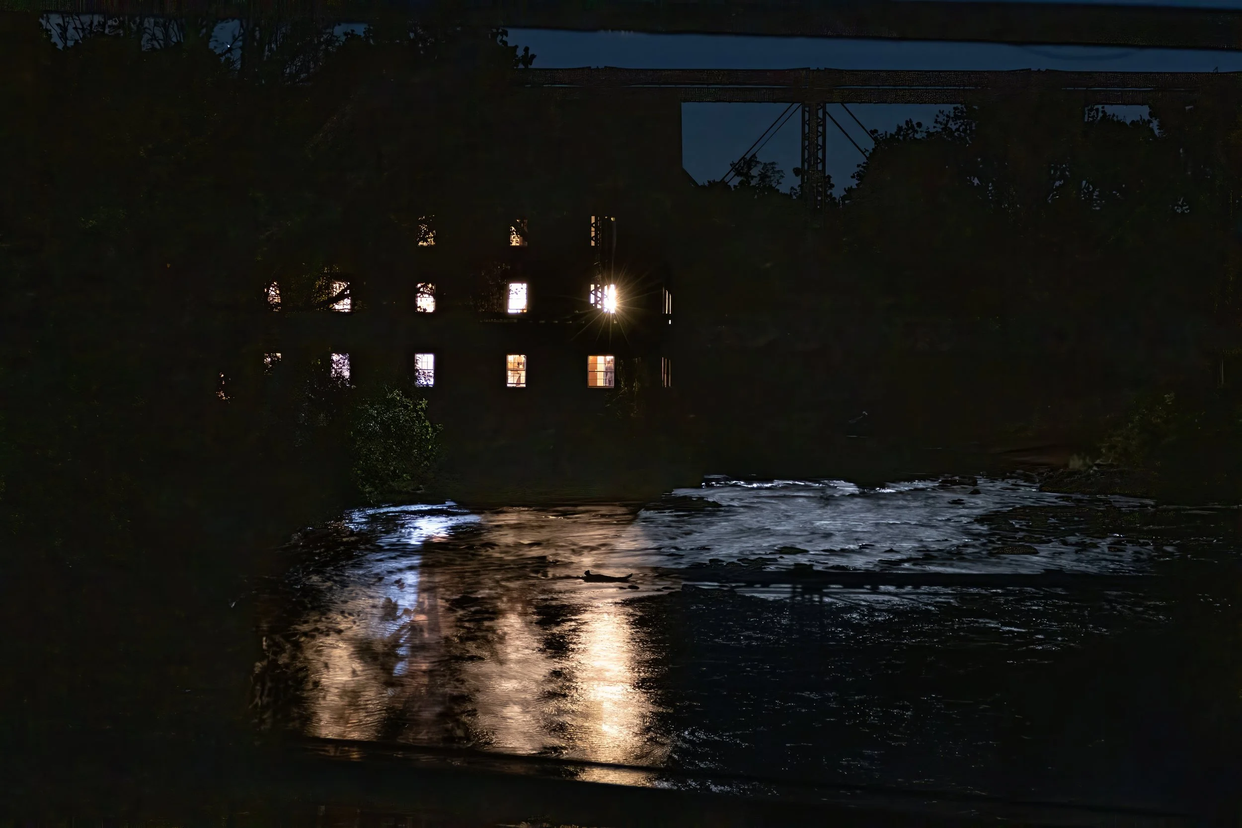 Nighttime scene of a factory or mill beside a river with illuminated windows, with reflections on the water surface.