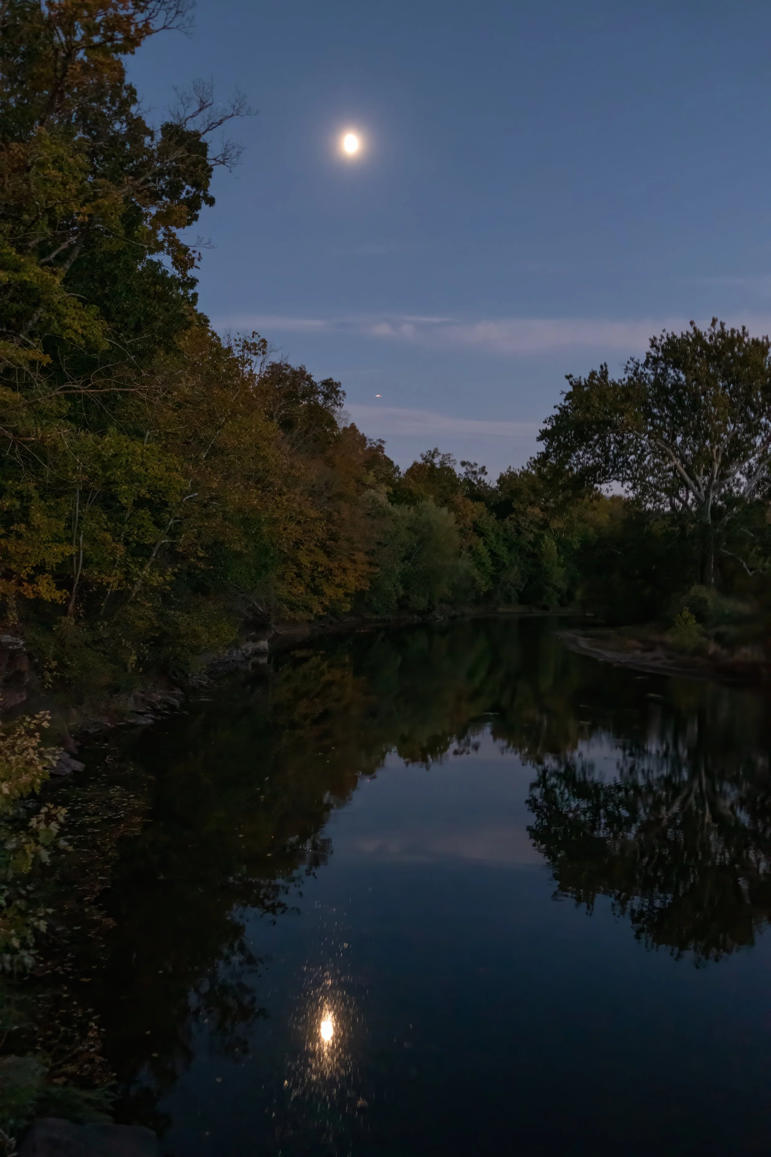Nighttime scene showing a river reflecting the moon and trees along its banks, with a partly cloudy sky.