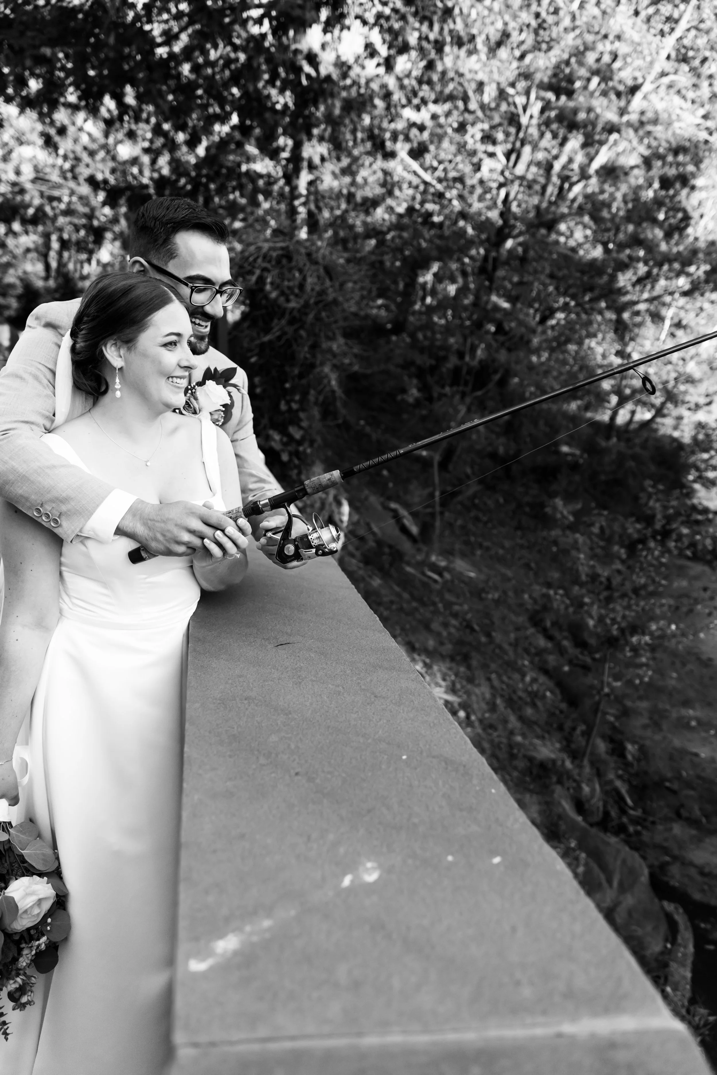 A smiling bride and groom stand together on a bridge, fishing by the South Branch Raritan River in Hillsborough, NJ, within a rural setting.