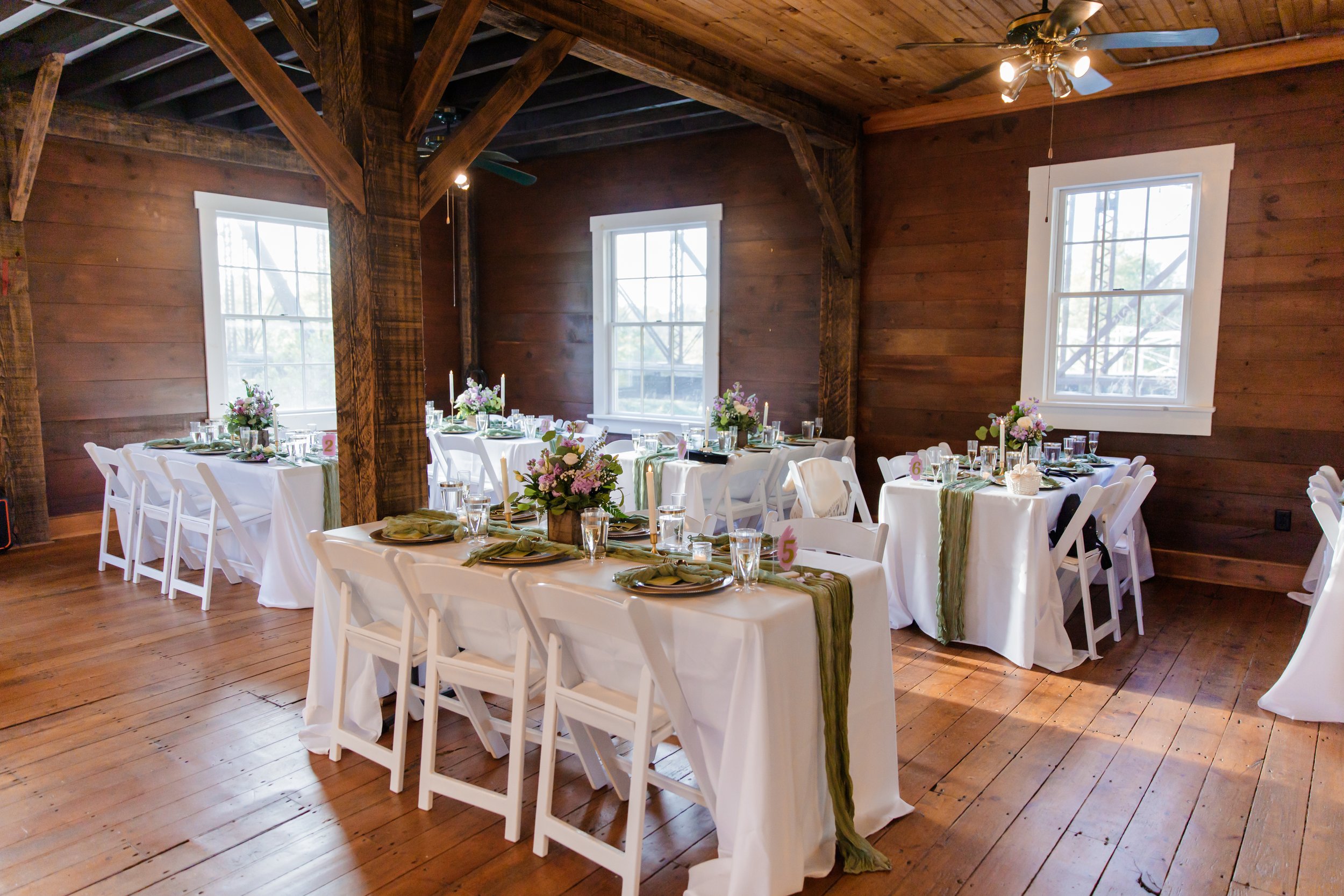 Decorated banquet hall with several round and rectangular tables covered with white tablecloths, green runners, and floral centerpieces, set with plates, glasses, and silverware, in a rustic wooden room with large windows and a ceiling fan.