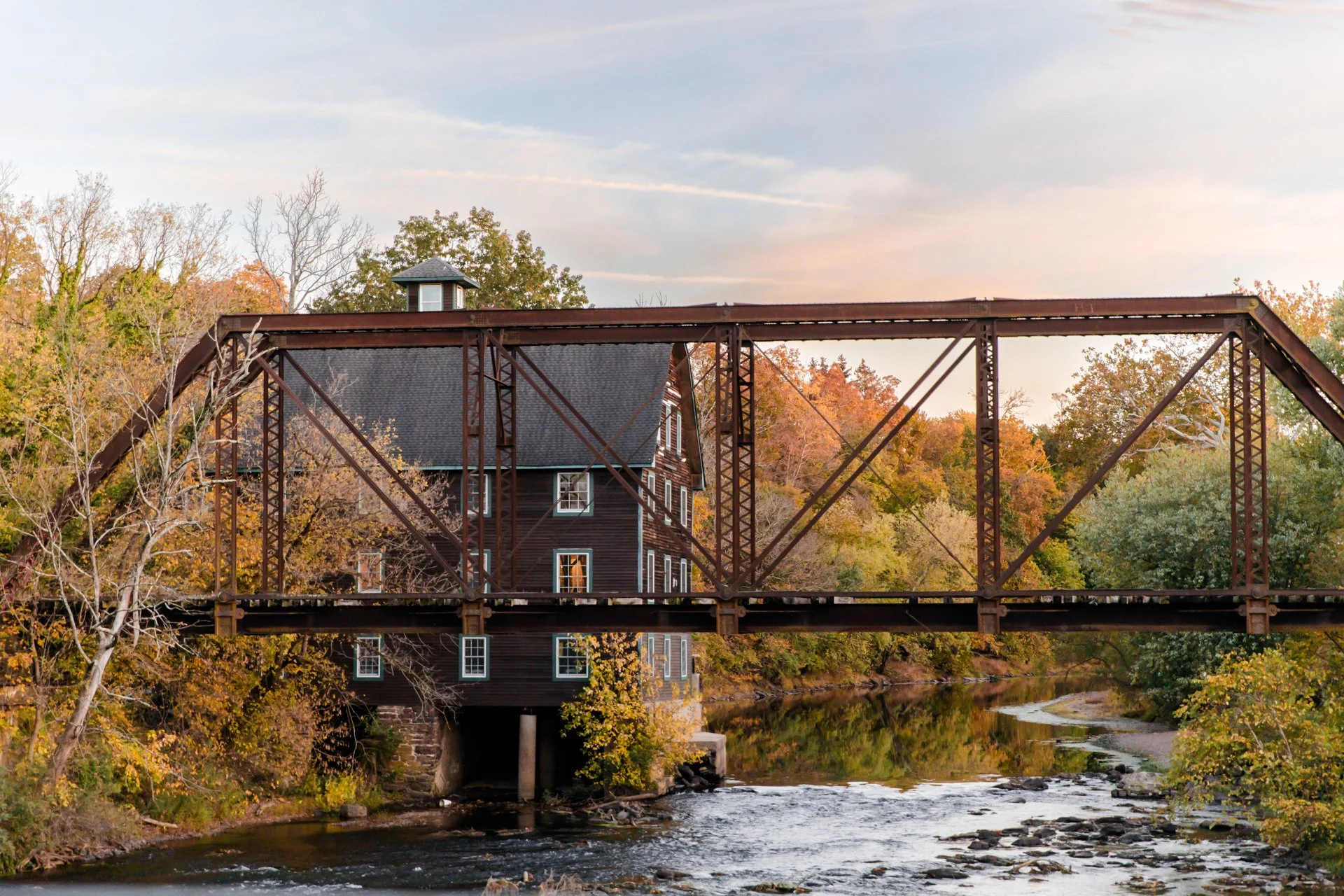 A historic wooden riverside mill in Neshanic Station, NJ. A rustic metal bridge crosses over a flowing river, surrounded by trees with autumn foliage.