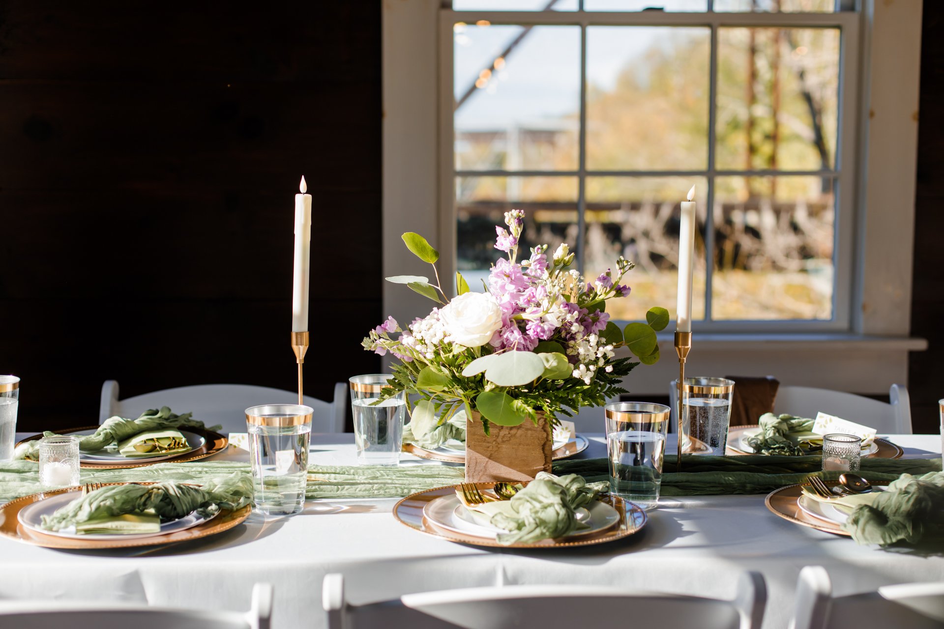 A beautifully set dining table indoors with a floral centerpiece, candles, glassware, and place settings. A window shows an outdoor scene with trees and a bridge in the background.