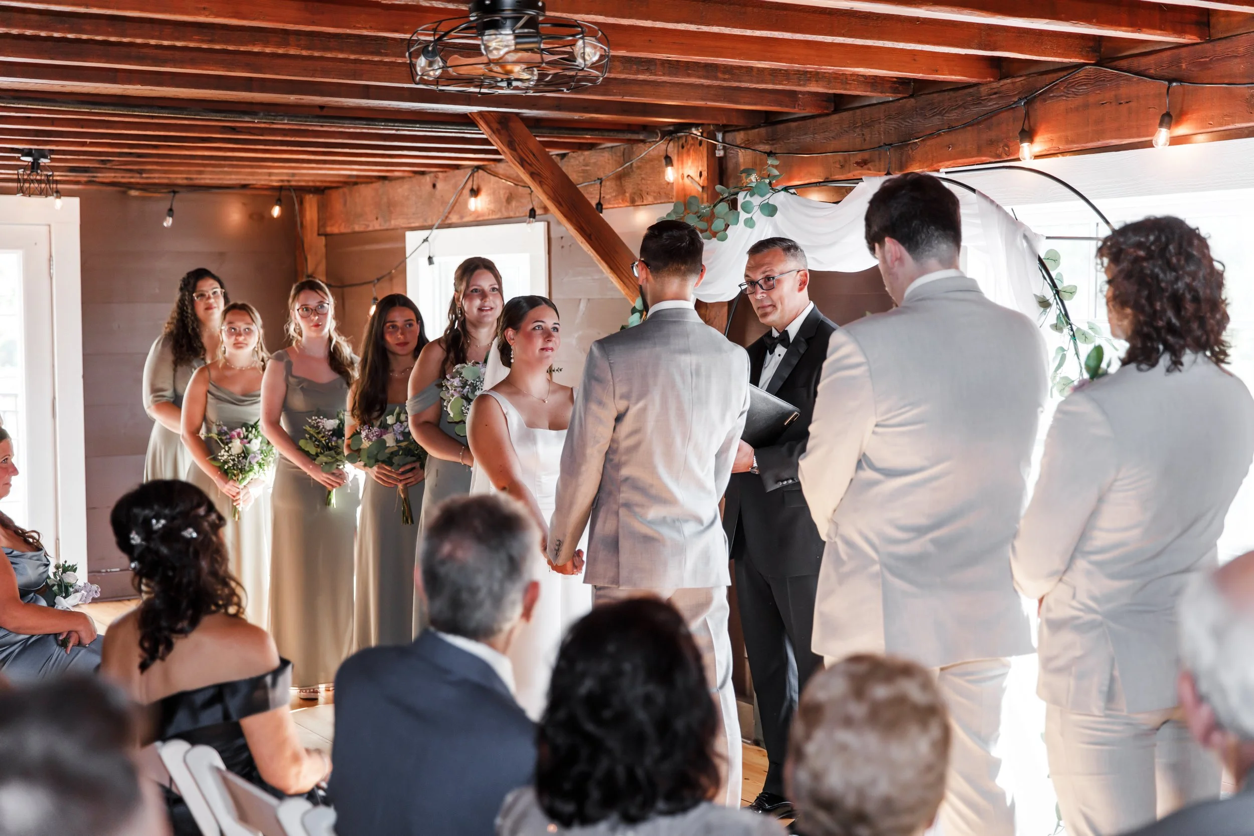 A wedding ceremony with the bride and groom holding hands facing each other, officiant reading from a book, bridesmaids standing in line holding bouquets, and guests seated watching inside a decorated wooden venue with string lights.
