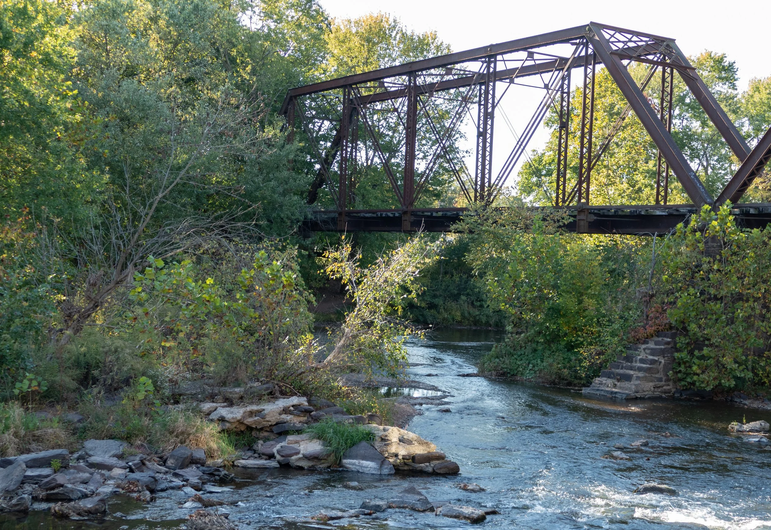 An old, rustic metal bridge in Neshanic Station, NJ over the South Branch Raritan River surrounded by green trees and rocks.