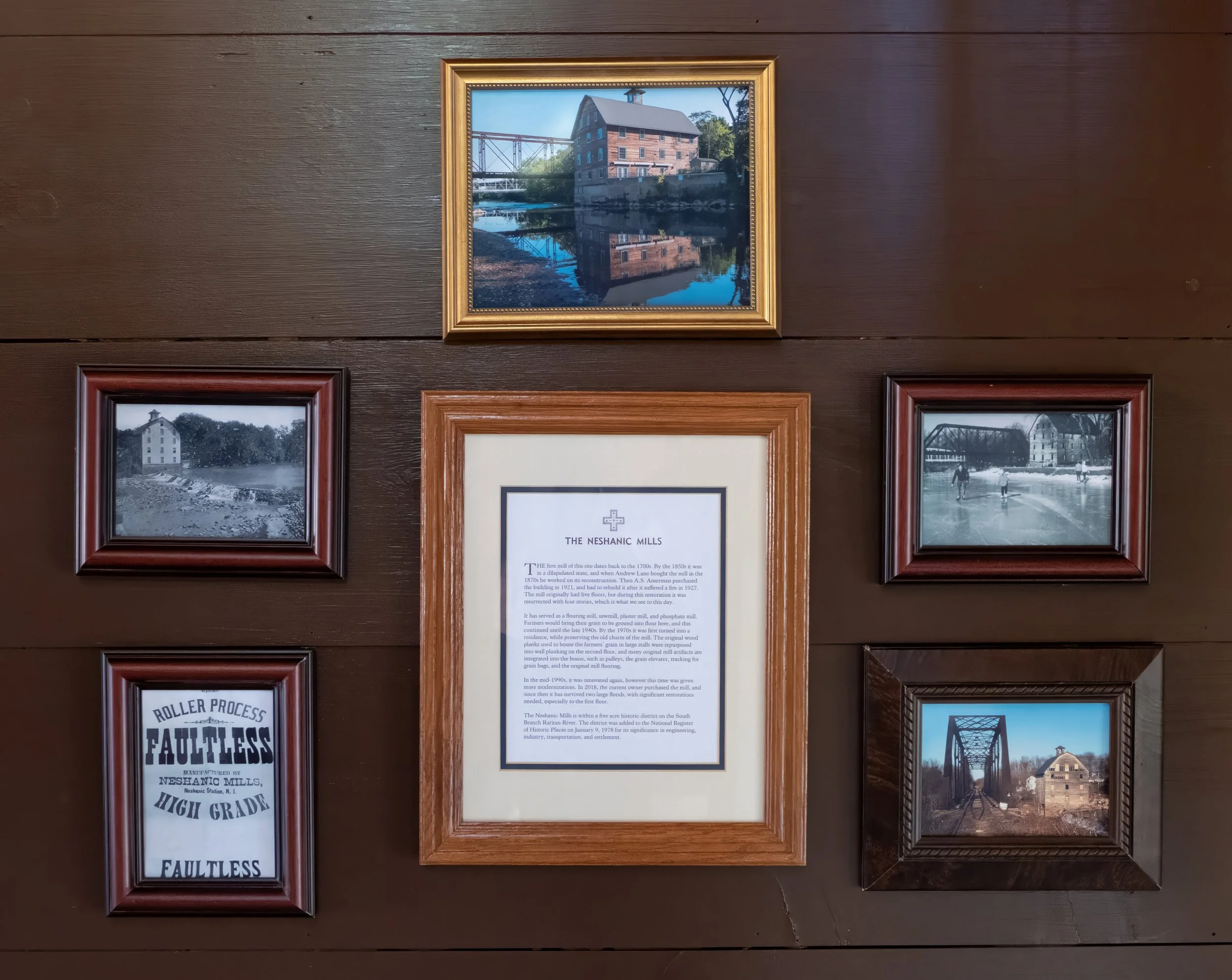 A wooden wall with six framed pictures and a text display, including an aerial view of a historic mill, black and white photos of old mills, an informational plaque about the Neshanic Mills, and a picture of a train bridge over a creek.