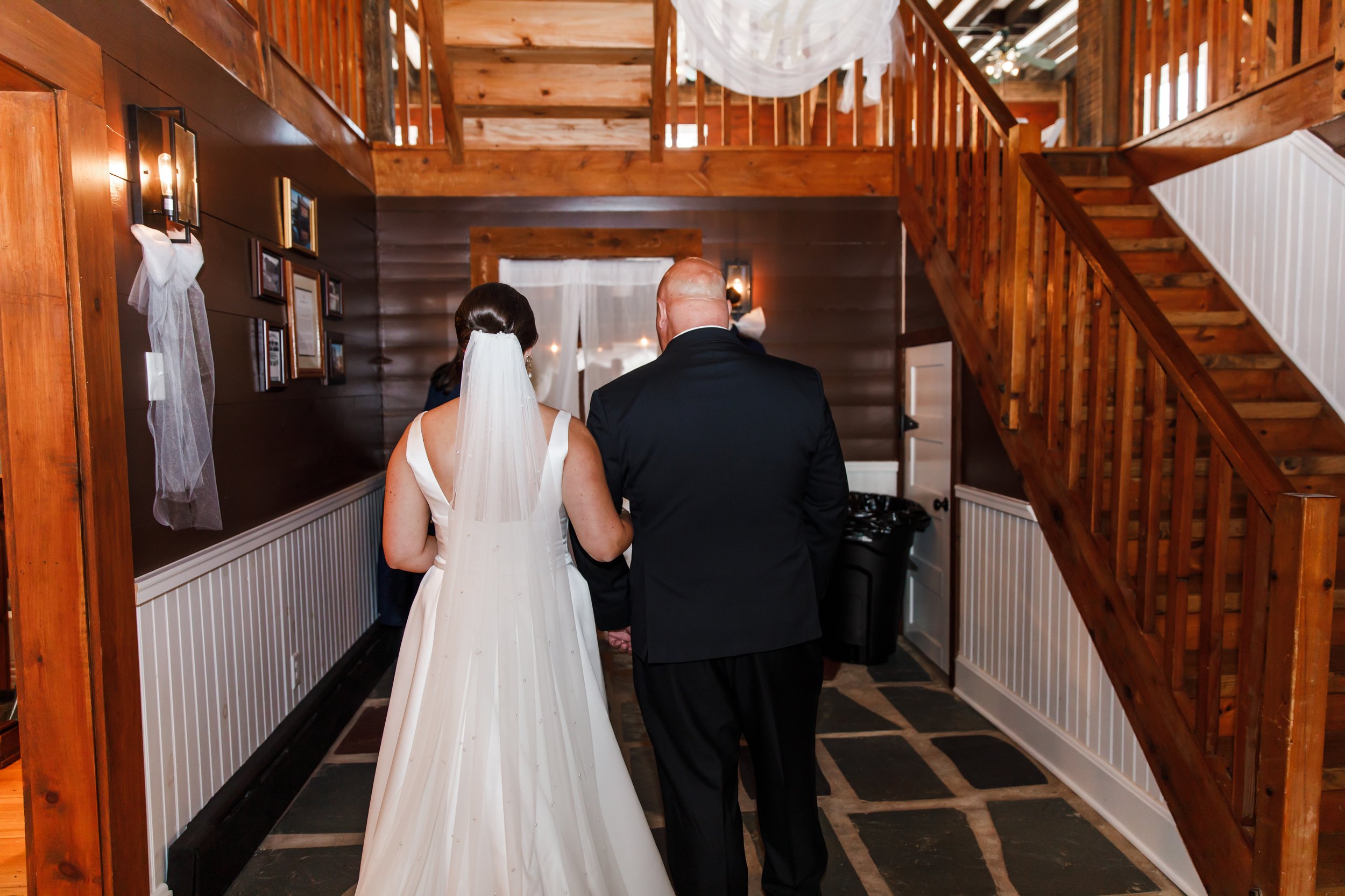 Bride and her father waiting to be introduced for her wedding inside a historic wooden venue, holding hands, with wooden stairs and wall decor visible.