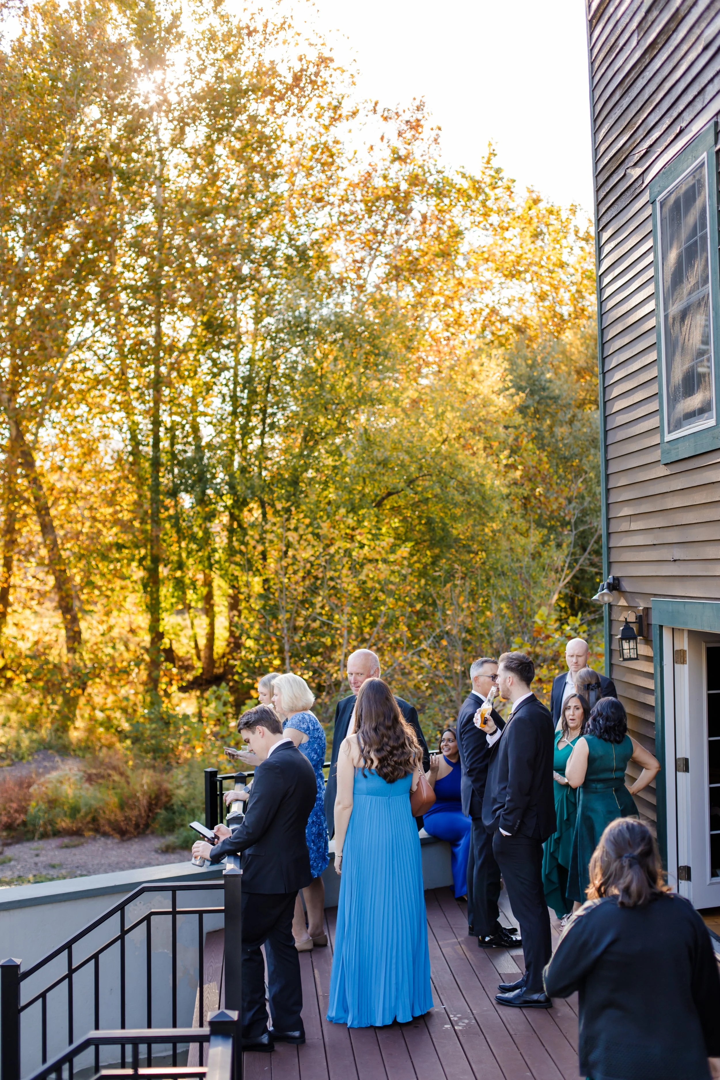 People dressed in formal attire gathering outdoors on a deck for a wedding cocktail hour in the fall, with trees in fall foliage in the background.