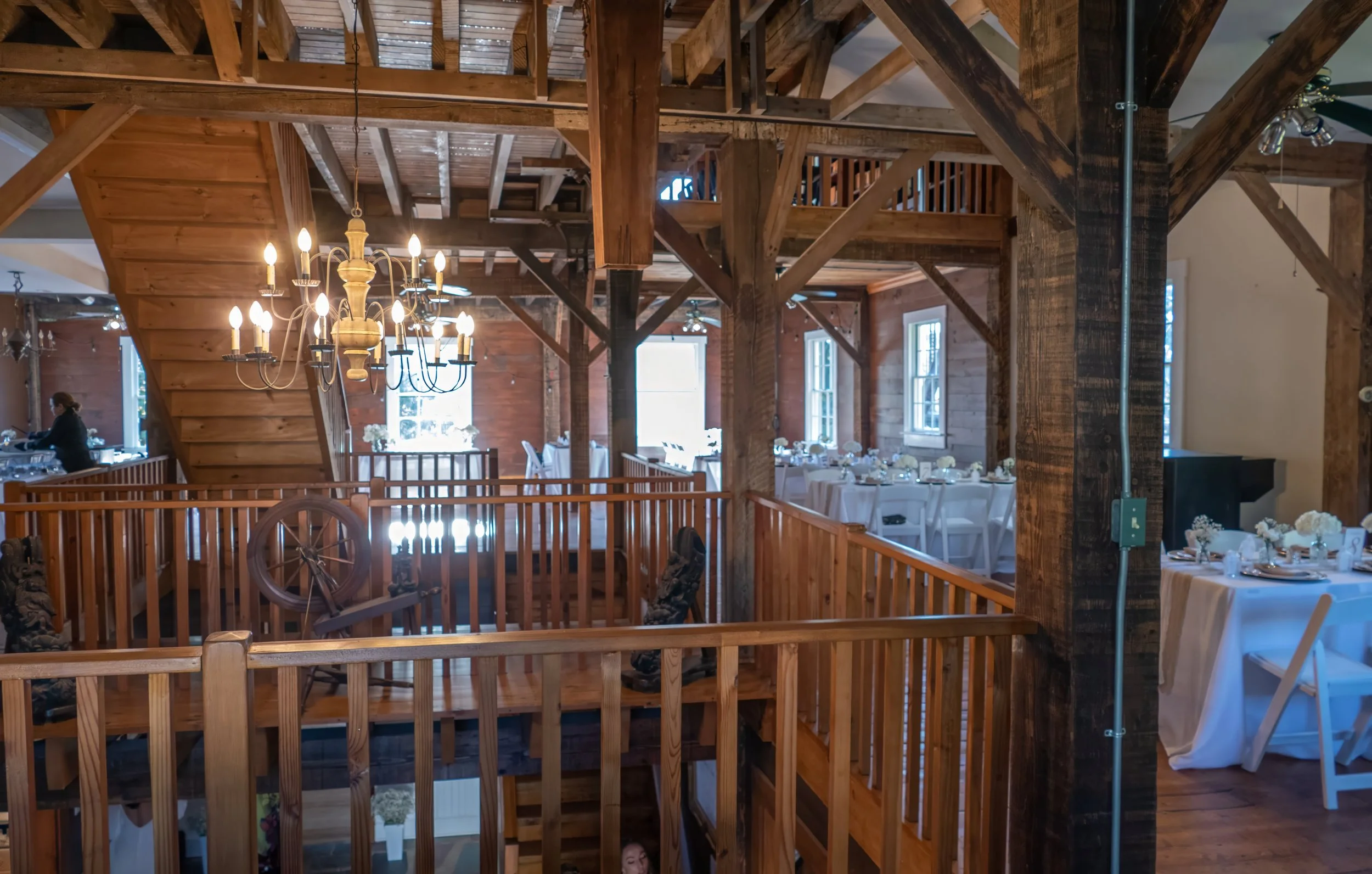 Interior of a rustic wooden venue decorated for a formal event with tables and chairs, chandelier lighting, and natural light from windows.