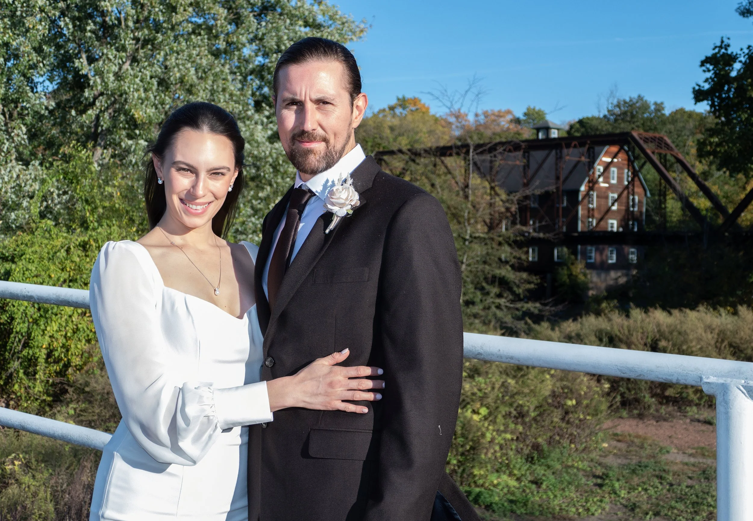 A smiling woman in a white wedding dress and a man in a black suit pose outdoors with a background of green trees, a blue sky, and The Neshanic Mill on the riverside.