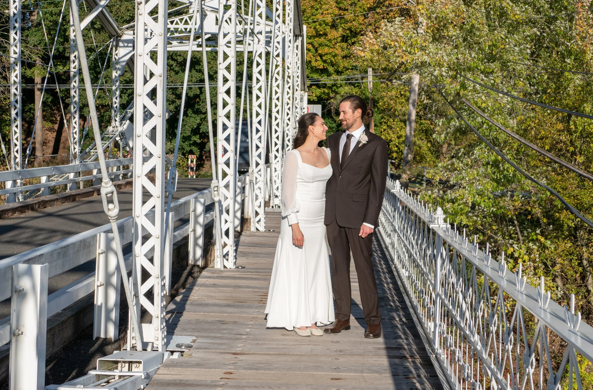 A newlywed couple in wedding attire standing on the Neshanic Station truss bridge, gazing at each other, with trees in the background during daytime.